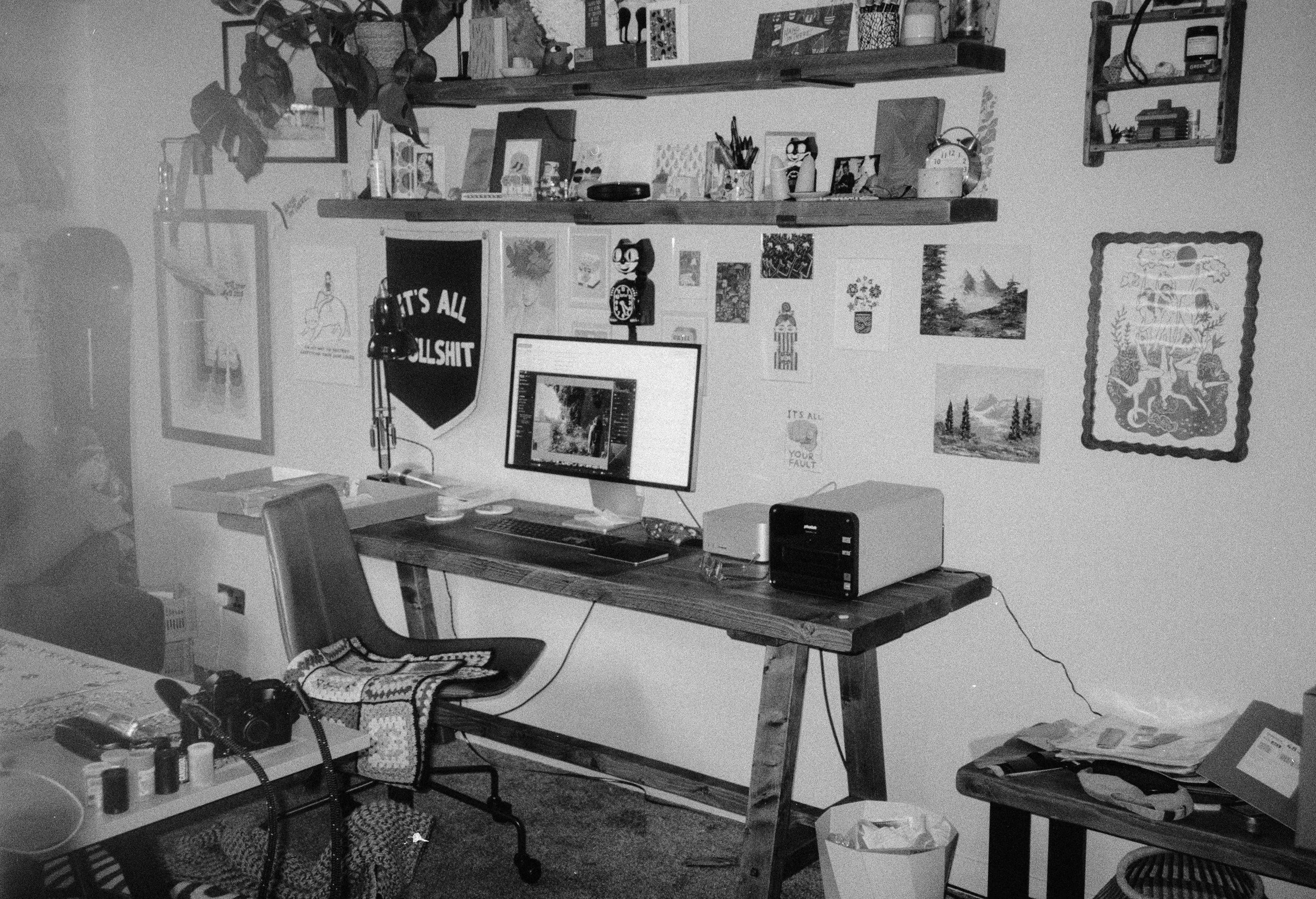 A cluttered home office with a desk, computer monitor, shelves filled with art supplies and decorations, and various items scattered around, including shoes, cups, and boxes.