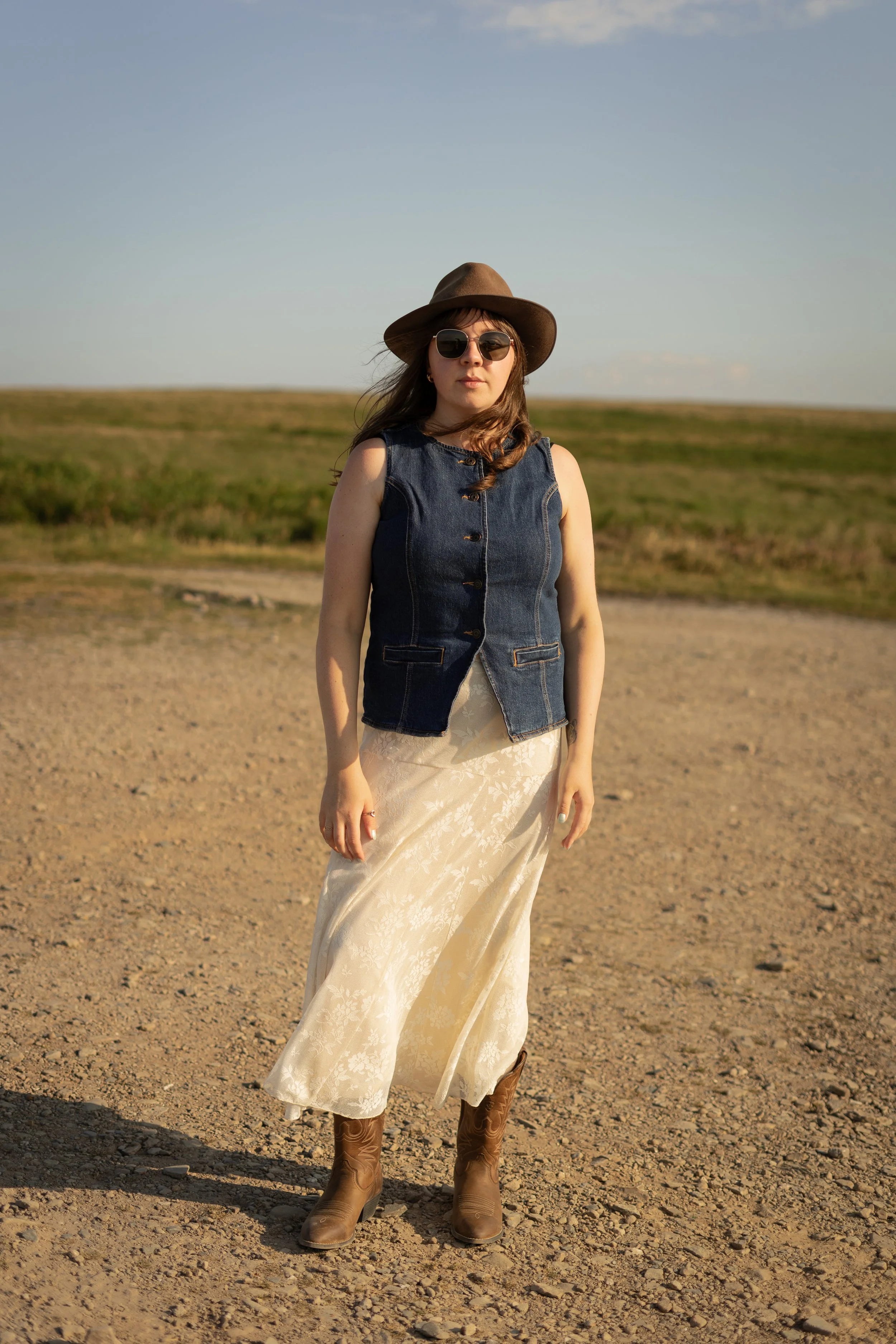 woman standing in open green and gravel space looking at camera with sunglasses and a hat on