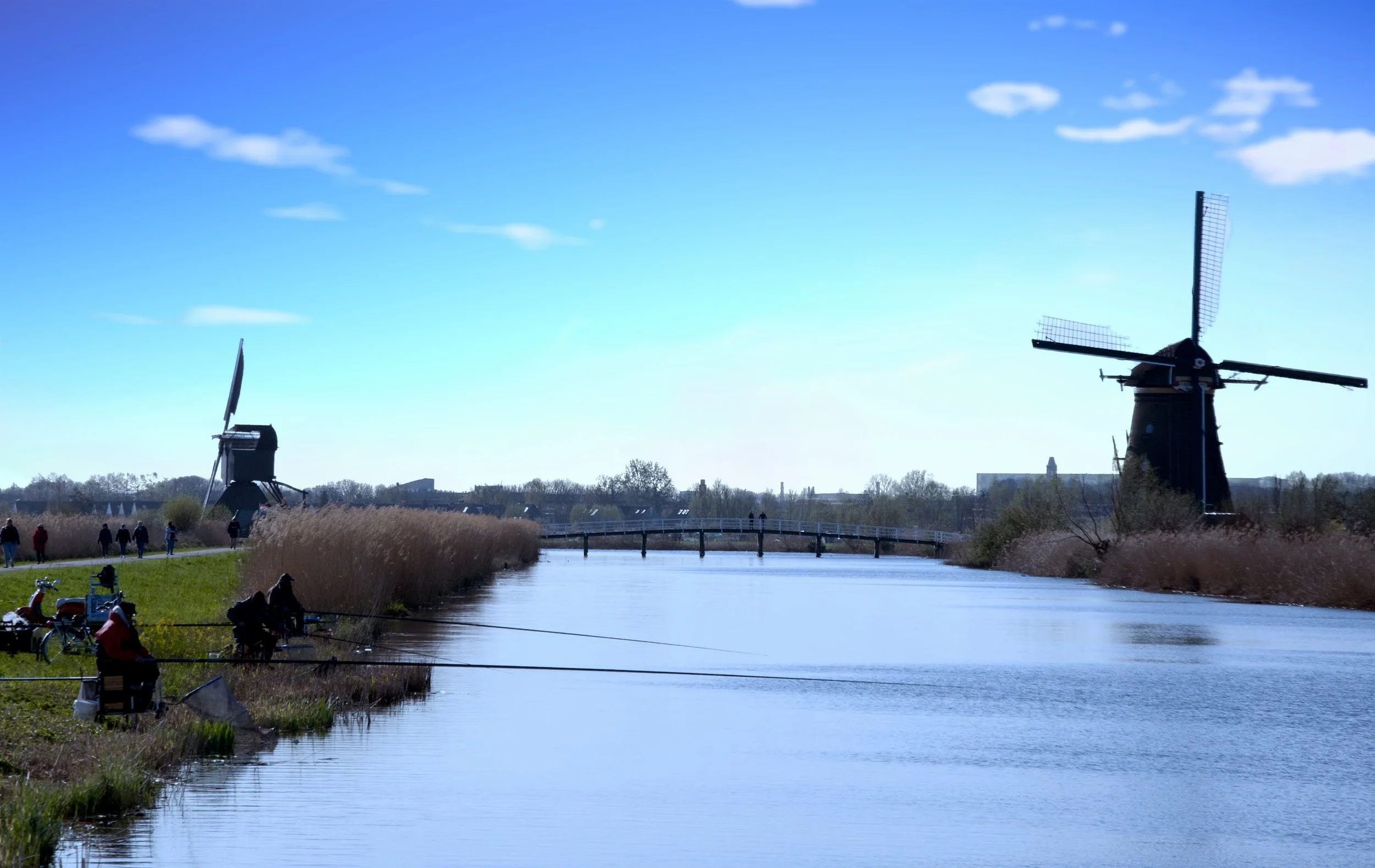 Two traditional Dutch windmills near a calm water canal, with people fishing and walking along the grassy banks on a clear, sunny day.