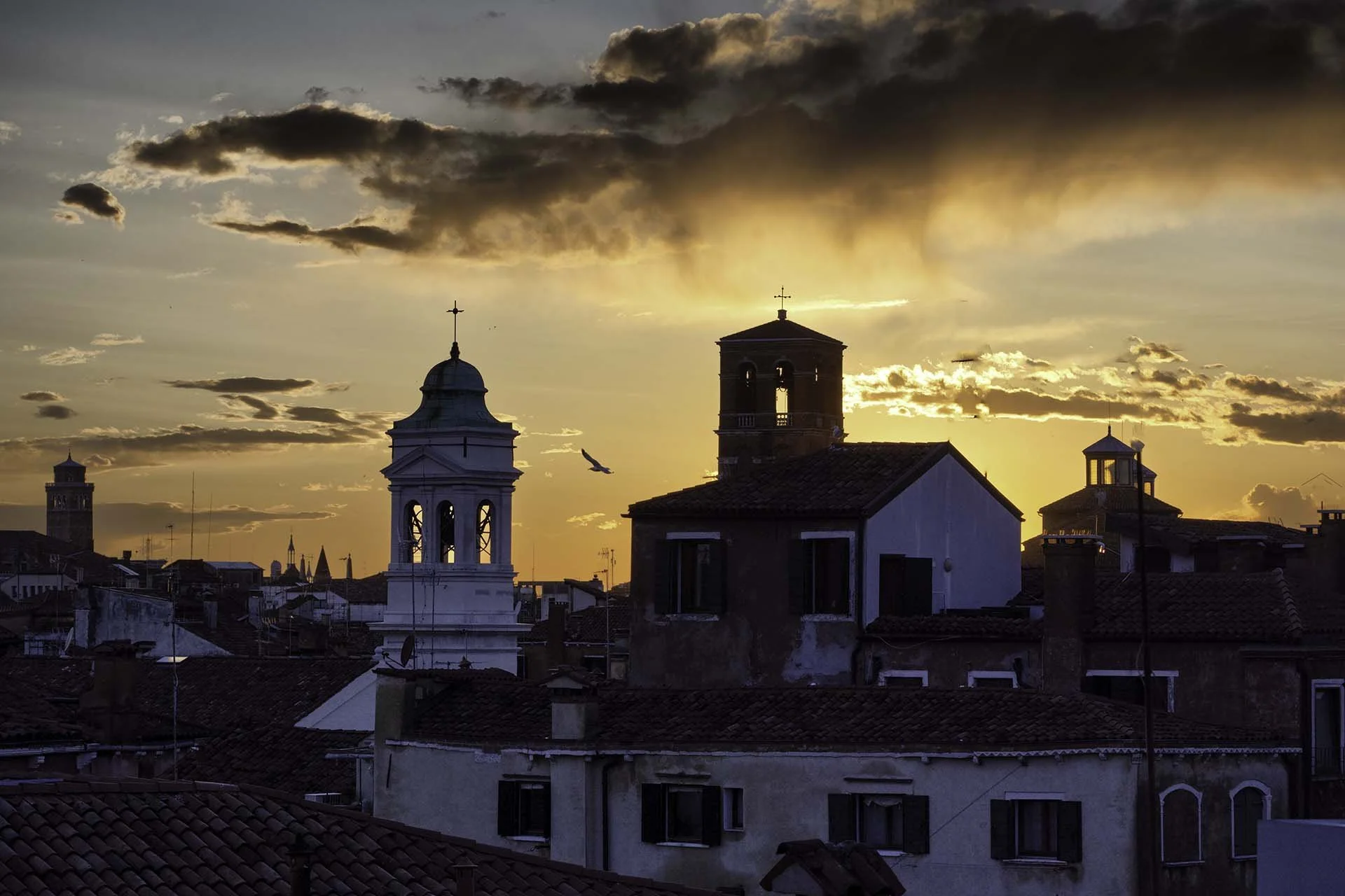 Sunset over a city with church steeples and rooftops, dark clouds in the sky with orange and yellow hues.