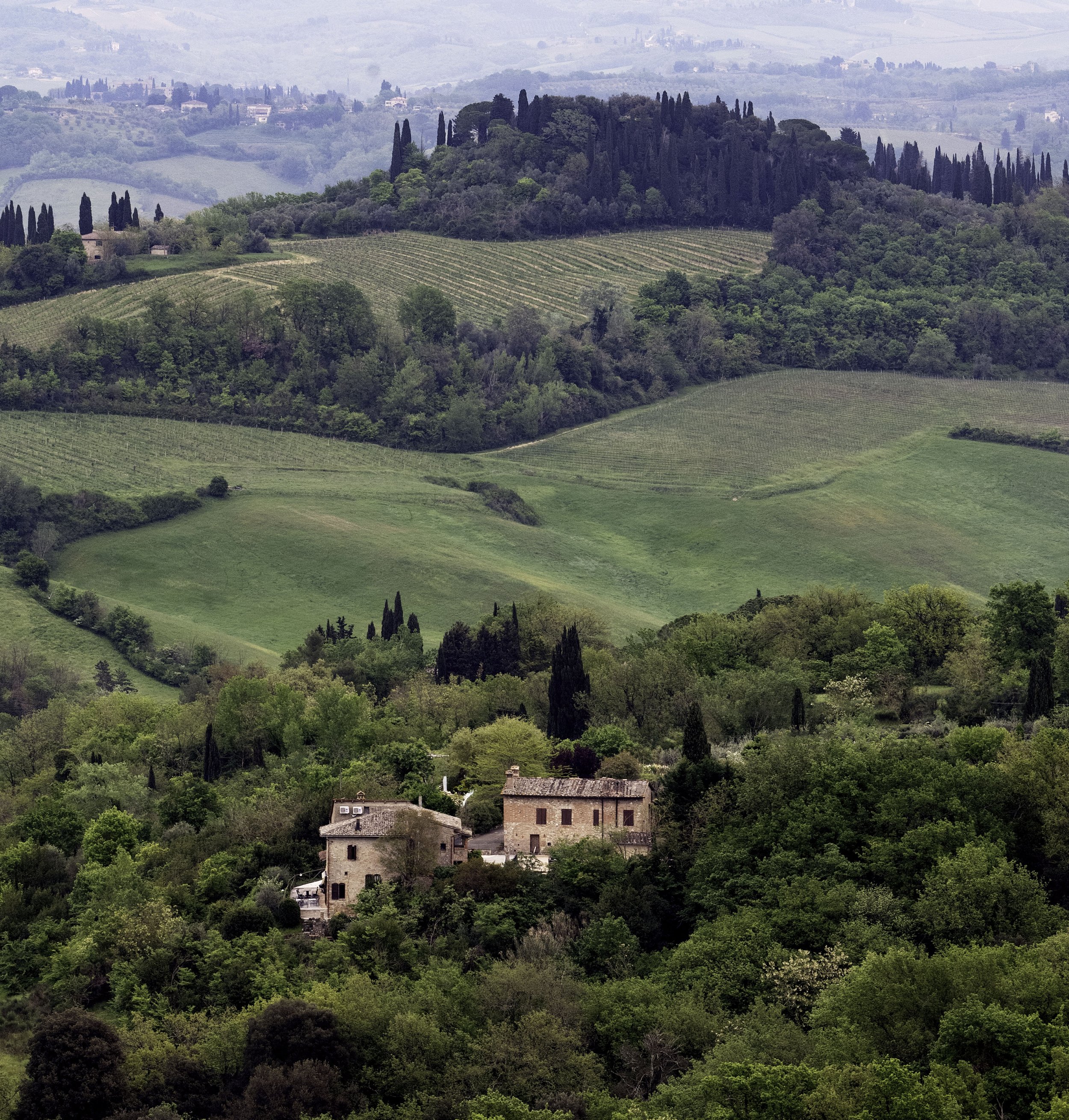 Scenic view of rolling green hills with dense trees and a small rustic house in the foreground, and distant countryside with more hills and buildings in the background.