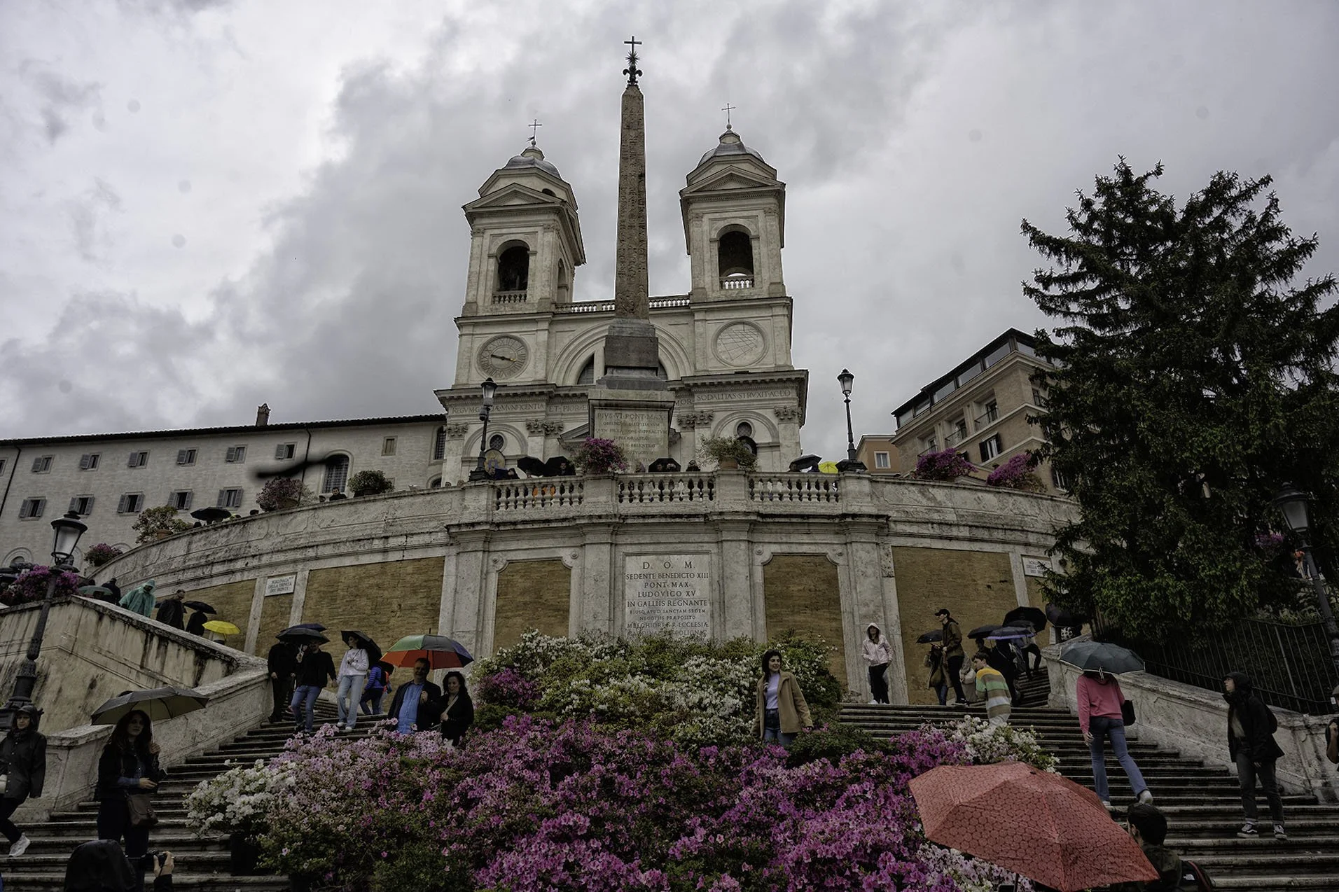 People walking down Roman-style steps decorated with pink and white flowers, umbrellas, and raincoats. Behind them, a white historic church with two bell towers and an obelisk in front, day overcast with cloudy sky.