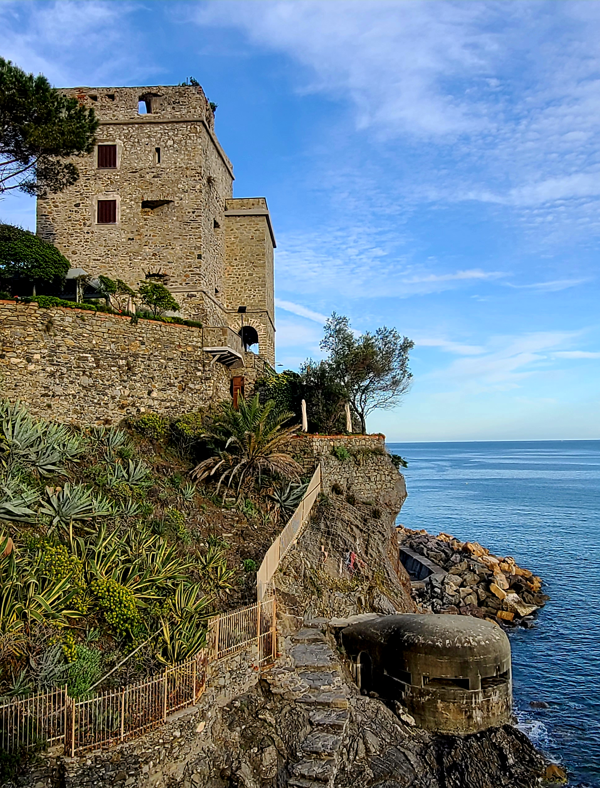 An old stone building with small windows is built on a hillside overlooking the ocean. There is vegetation and a pathway with a wooden fence, and rocks along the shoreline under a partly cloudy sky.