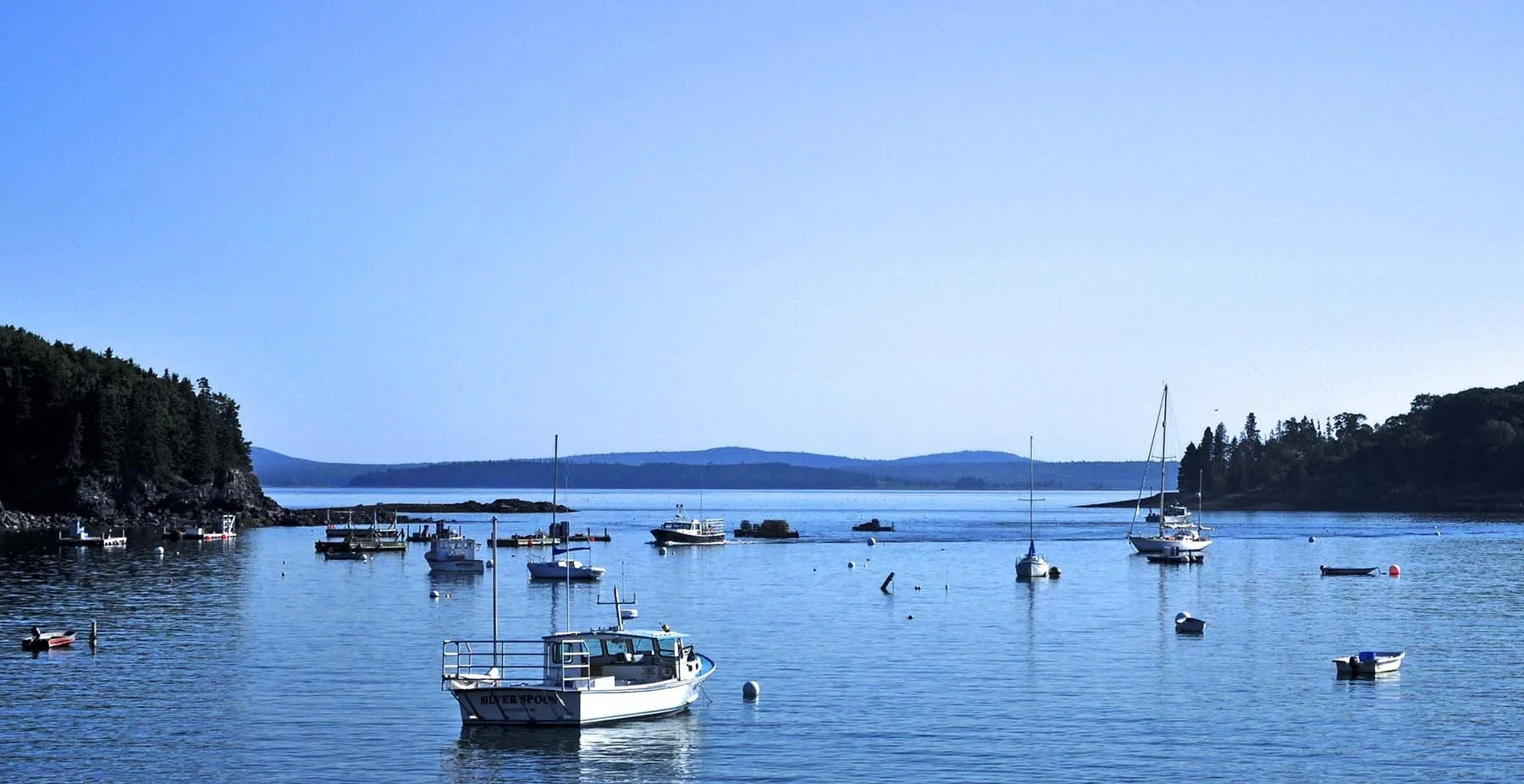 A calm harbor with several moored boats and sailboats, surrounded by green hills and a clear blue sky.