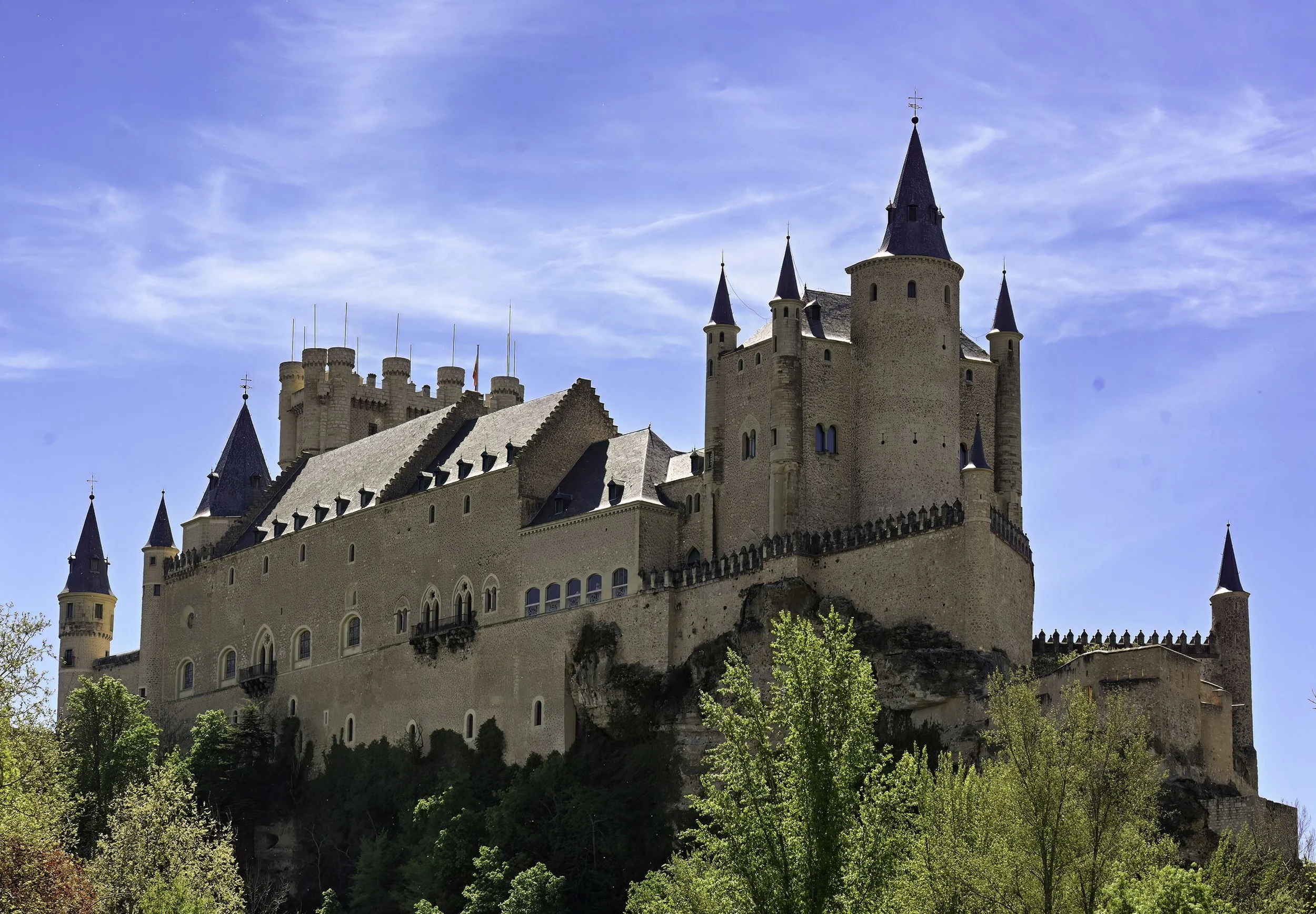 A large medieval castle with towers and turrets on a hillside, surrounded by green trees under a blue sky.