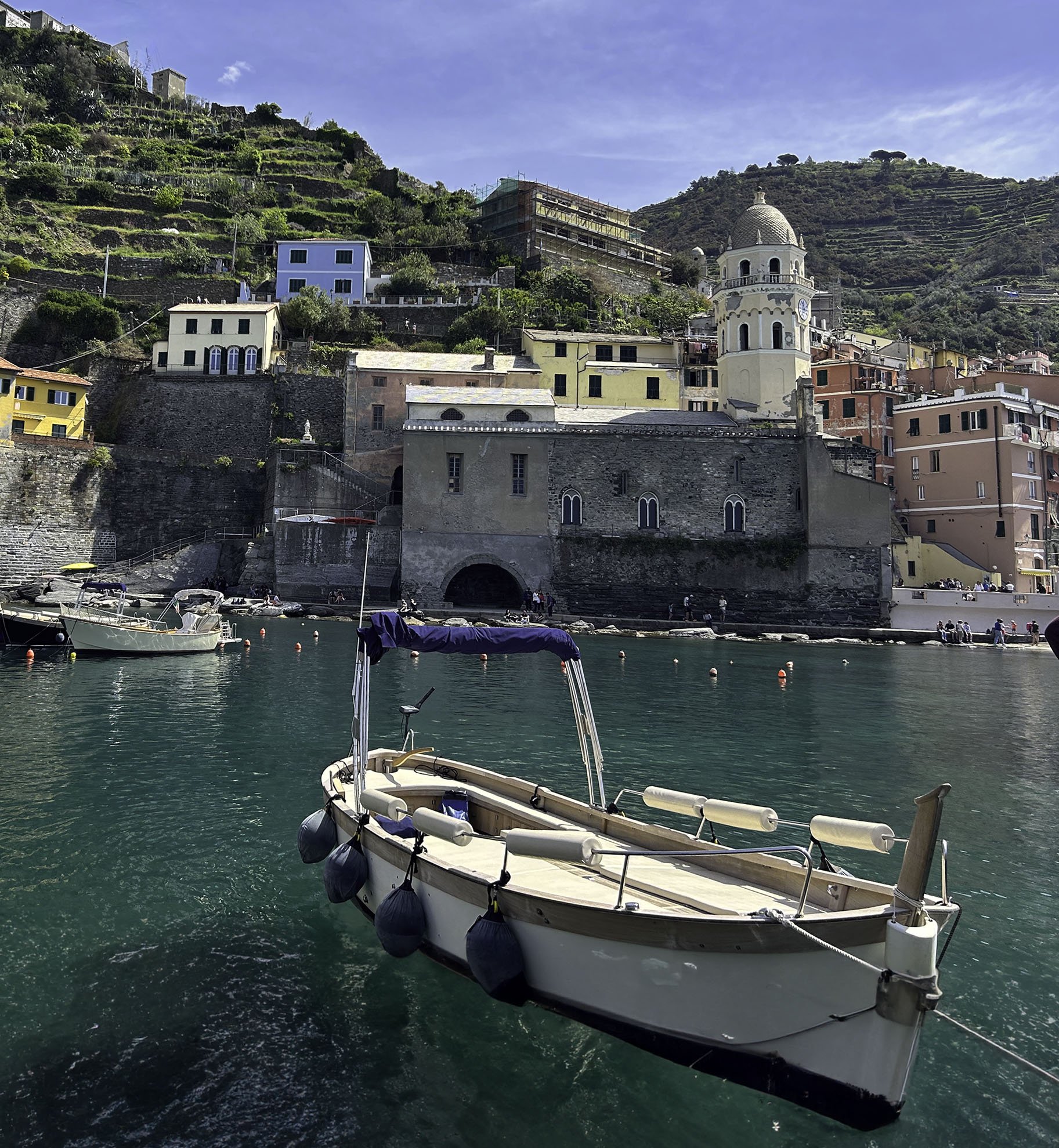 A small white motorboat with black fenders is docked in a harbor with greenish water. The boat is in front of a seaside town built on a hillside with colorful buildings and a clock tower. The hillside has terraced gardens and a clear blue sky above.