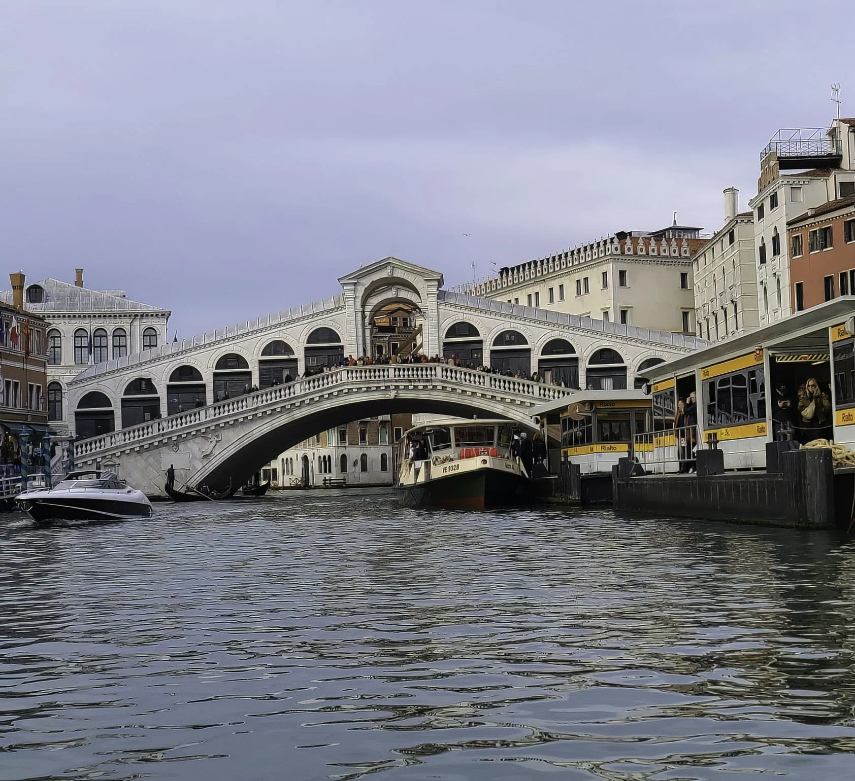 A white arched bridge over a canal with buildings on either side, a boat passing underneath, and a water bus docked at the canal side.