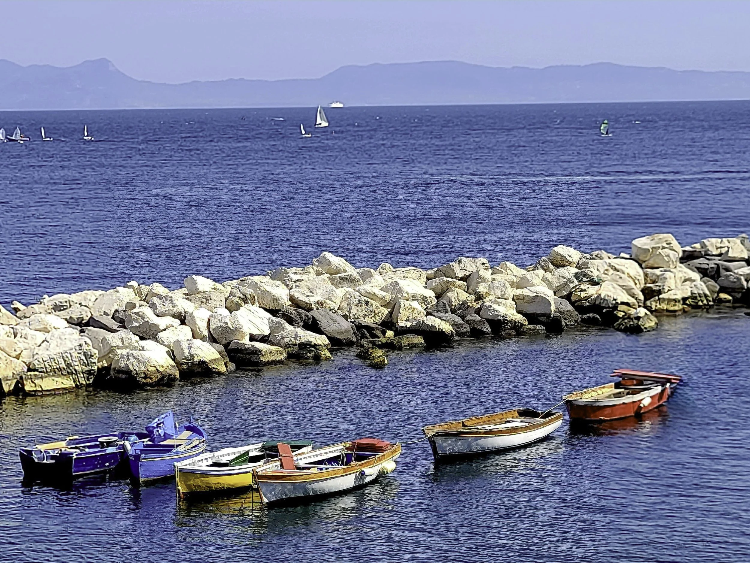 Several small colorful boats are docked near a rocky shoreline in a harbor, with a calm blue sea extending to a distant mountainous landscape.