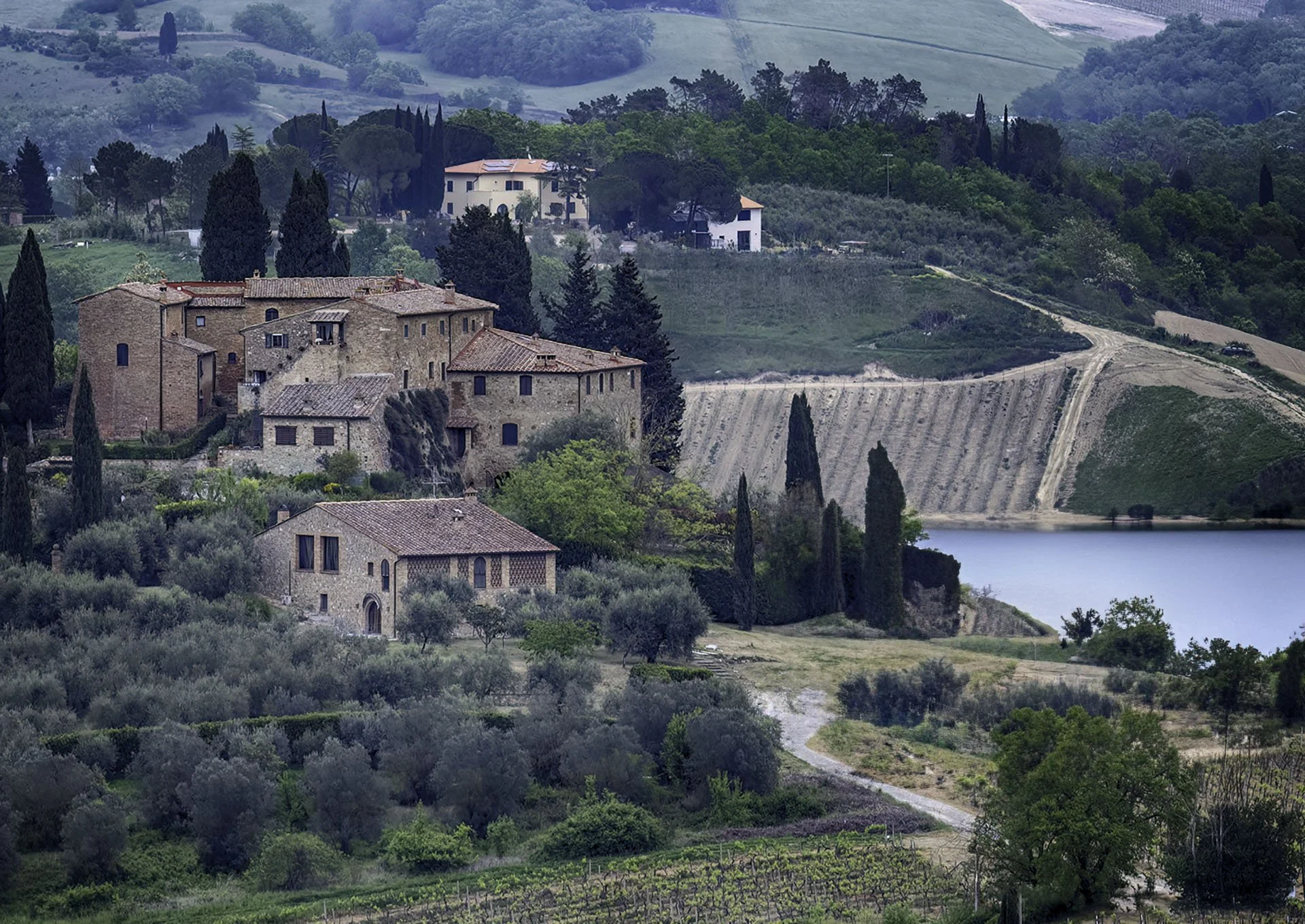 A scenic landscape of rolling green hills with a small lake in the foreground, featuring a historic stone building with a tiled roof, surrounded by tall cypress trees and lush vegetation.