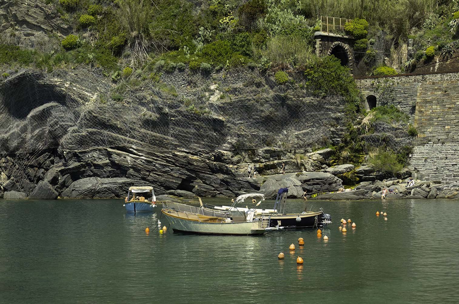 Boats anchored in a calm green lake near rocky shores with people sitting and walking on rocks, and a hillside with bushes, trees, and stone structures in the background.