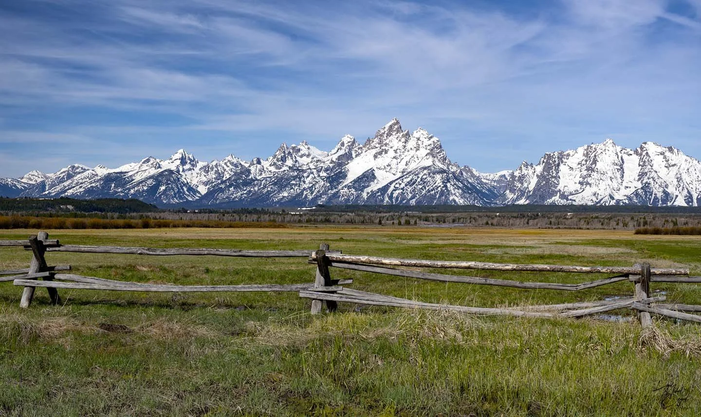Snow-capped mountains in the distance with a grassy field and rustic wooden fence in the foreground under a partly cloudy sky.