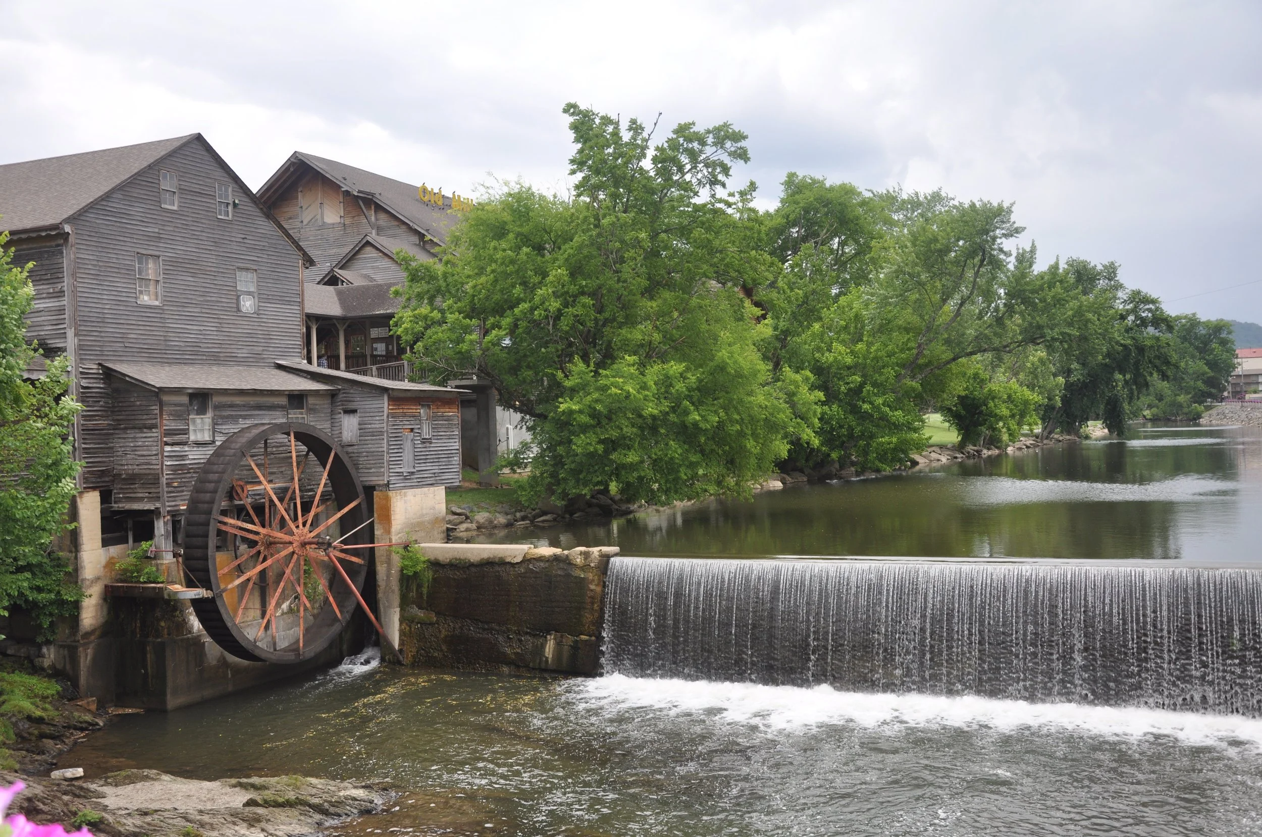 An old wooden grist mill by a river with a water wheel and lush green trees, with a cloudy sky overhead.