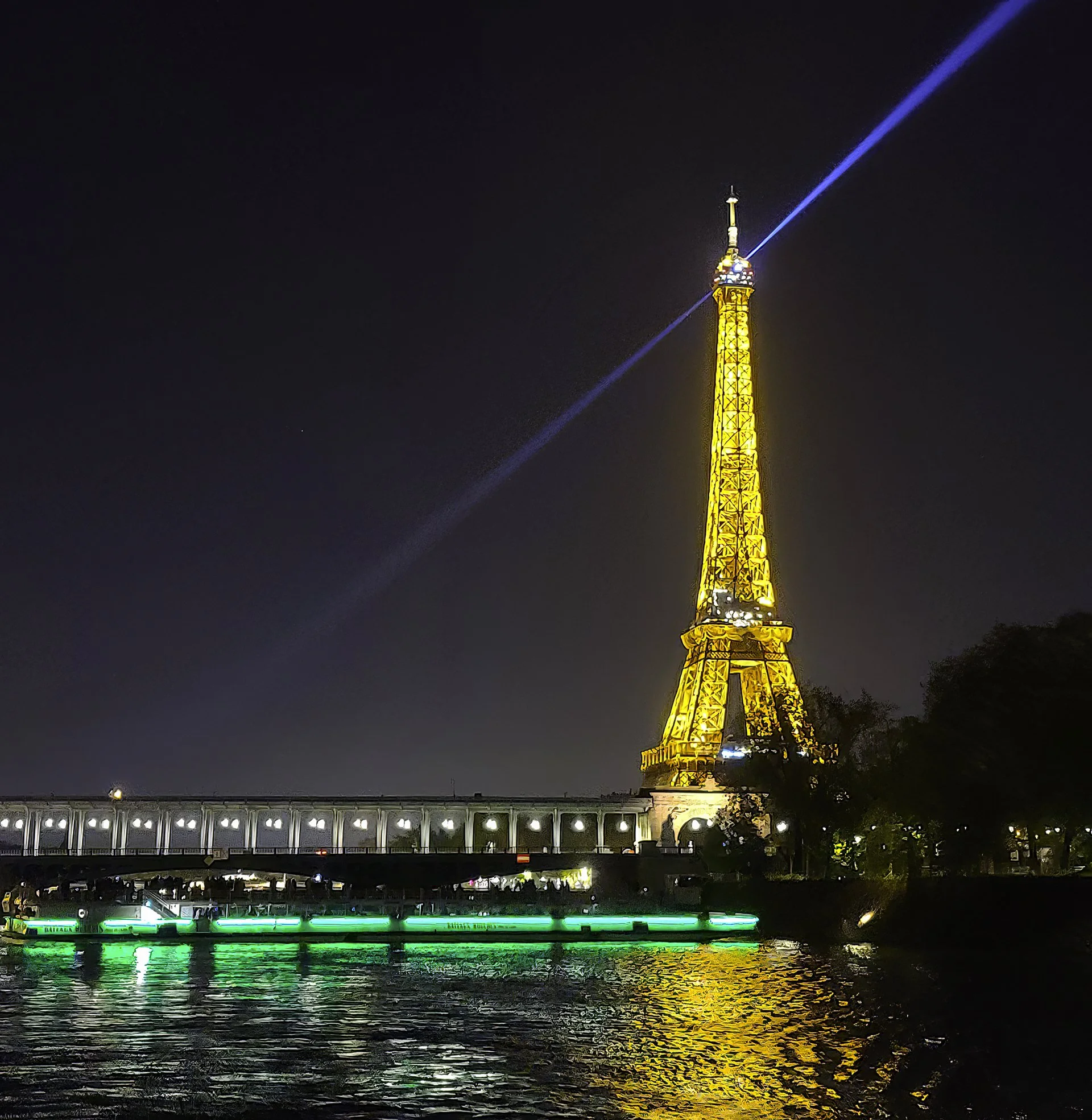 Night view of the Eiffel Tower in Paris illuminated in yellow with a blue beam of light at the top, reflections shimmering on the river below, and a bridge with green lights crossing the river in the foreground.
