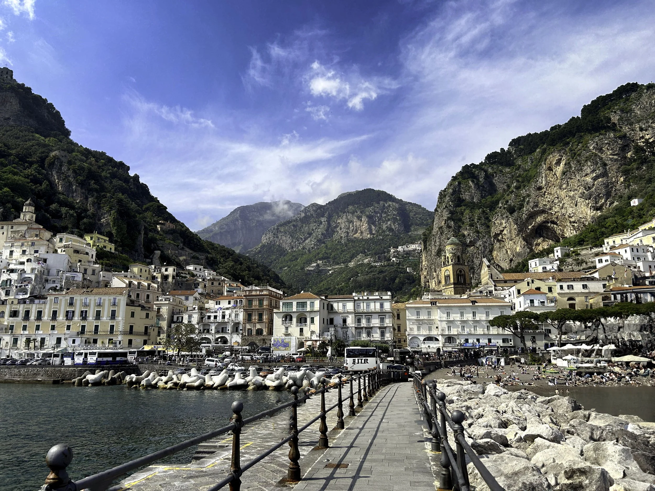 A coastal town nestled in a valley with multicolored buildings, surrounded by green mountains with clouds and blue sky overhead. A harbor with boats and a rocky beach with people sunbathing is in the foreground.