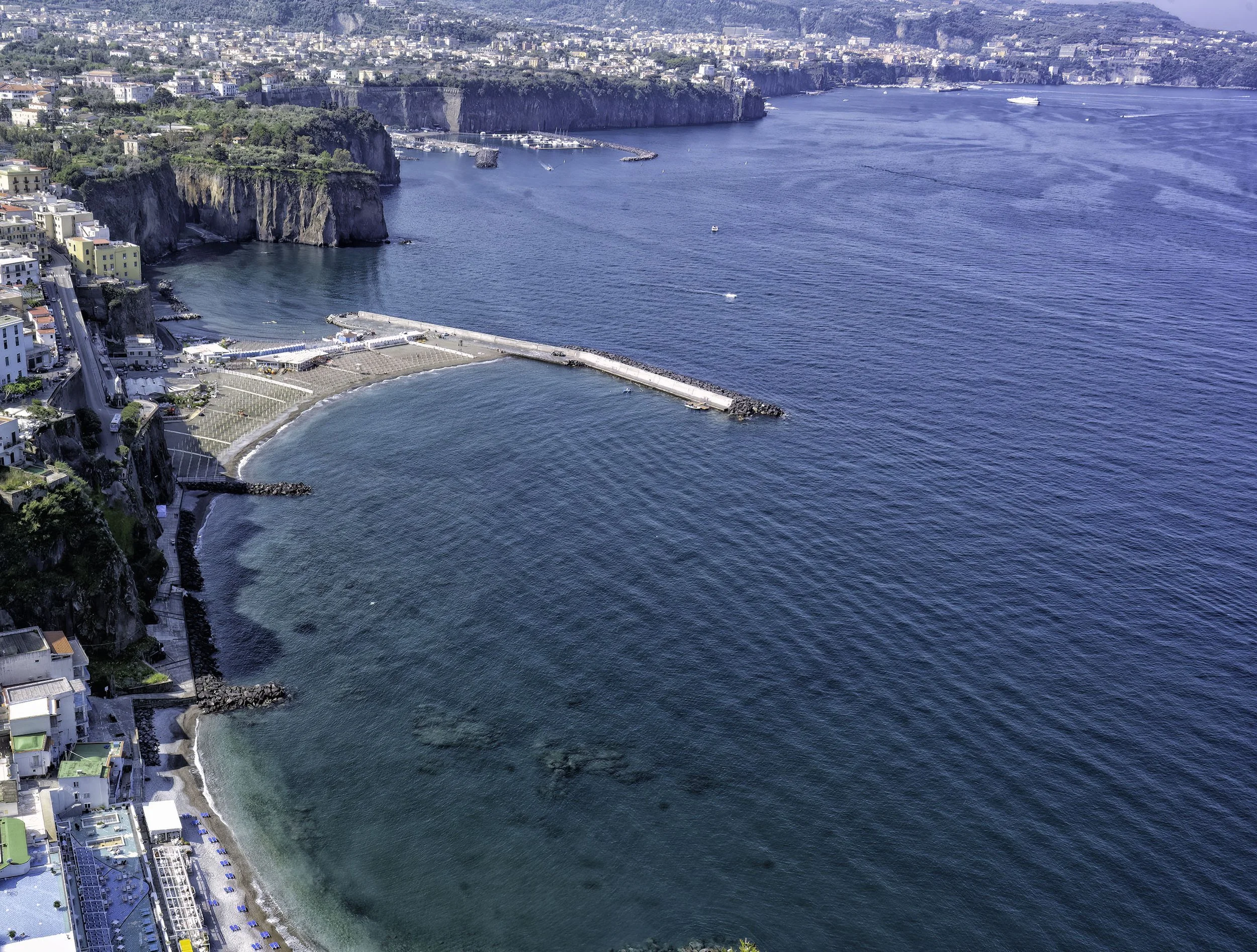 Aerial view of a coastal city with cliffs, a harbor with boats, and buildings along the shoreline.