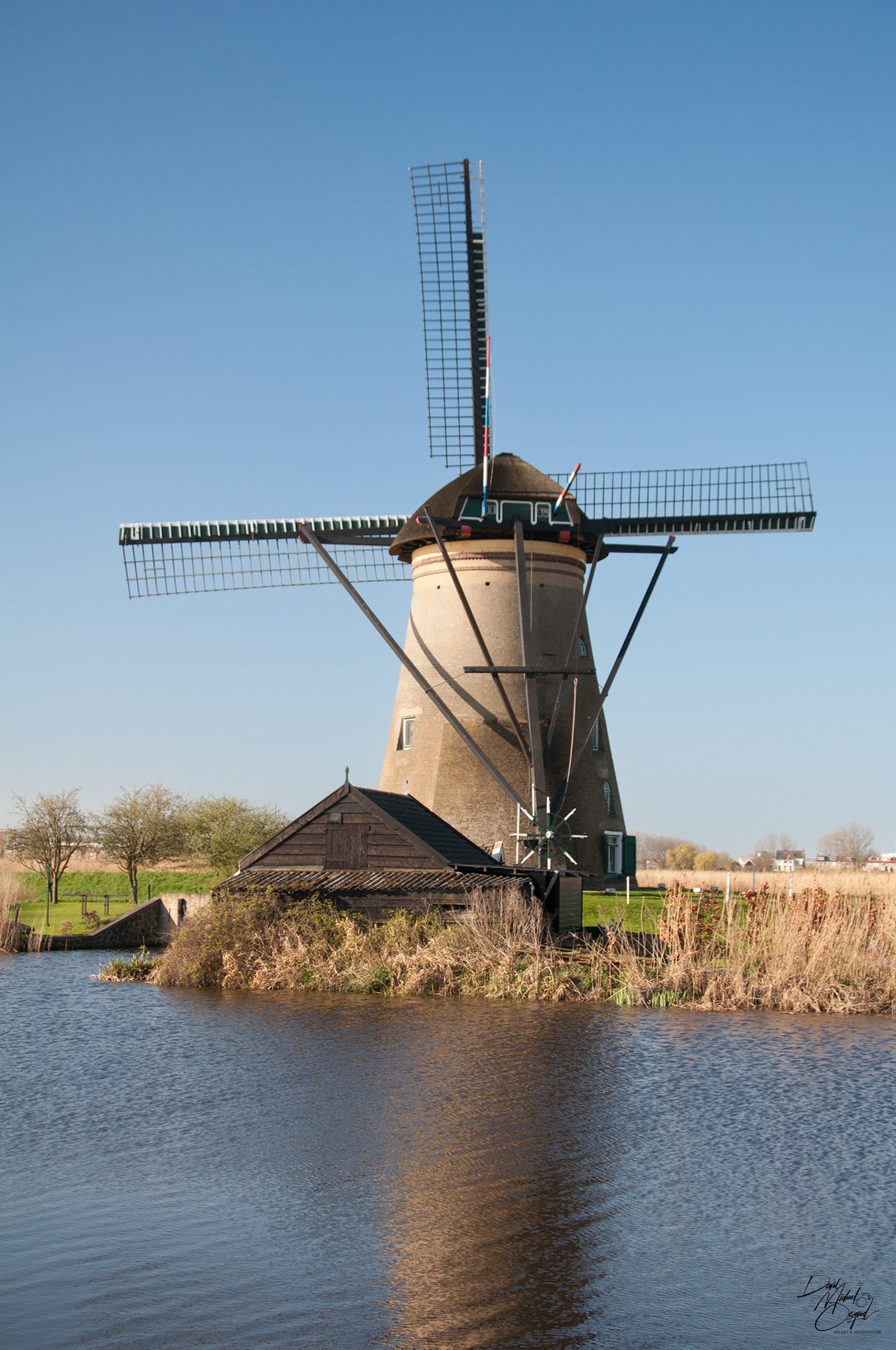 A traditional windmill with four large blades standing next to a body of water with trees and houses in the background under a clear blue sky.