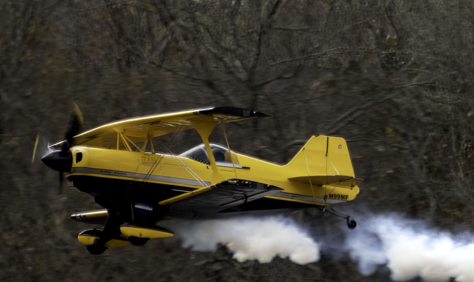 Yellow aerobatic airplane flying low over a mountainous terrain, leaving white smoke trail behind.