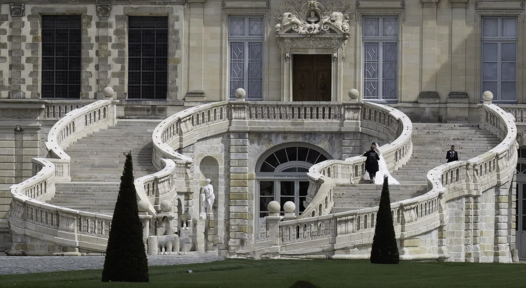 A grand staircase with intricate white railings and sculpted lion heads leading up to a large ornate building entrance with tall windows and detailed stonework. Two people, possibly a bride in a white dress and a groom or a man in formal attire, are 