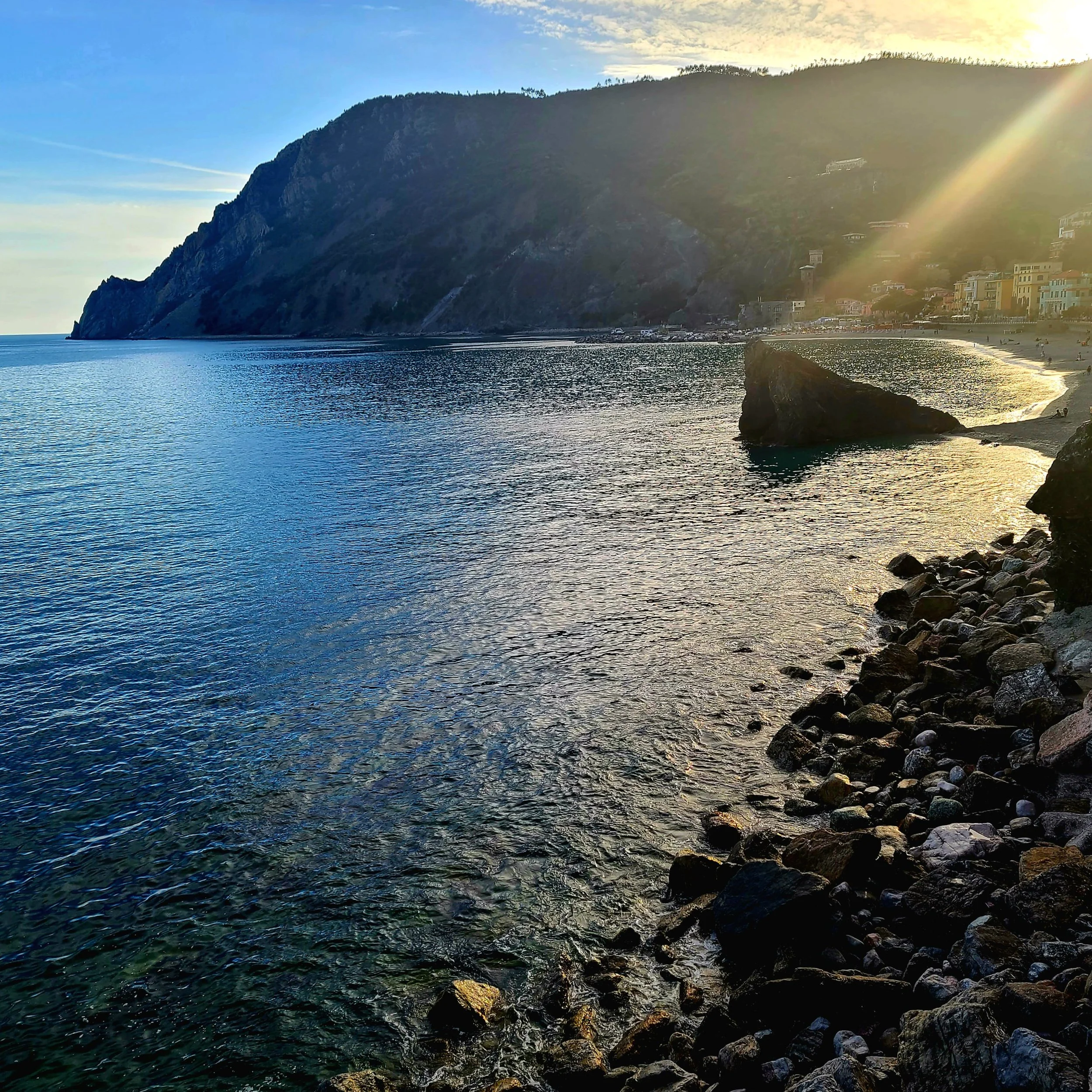 Sunset over a coastal hillside with buildings and a beach, calm water in the foreground, and a partly cloudy sky.