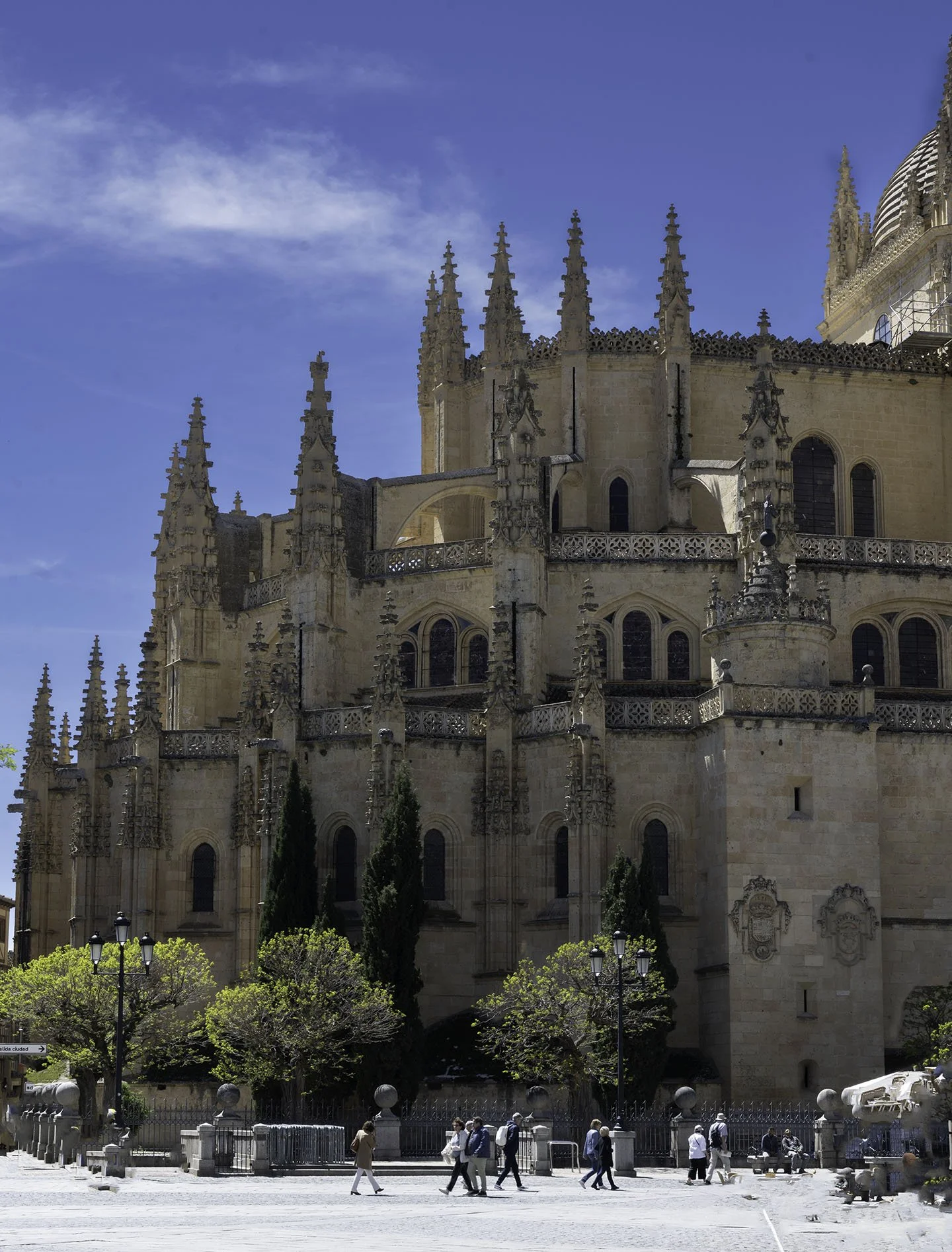 A historic cathedral with ornate Gothic architecture, pointed spires, and detailed stone carvings, set against a blue sky with some clouds. There are people walking in front of the cathedral, and trees and street lamps are visible in the foreground.