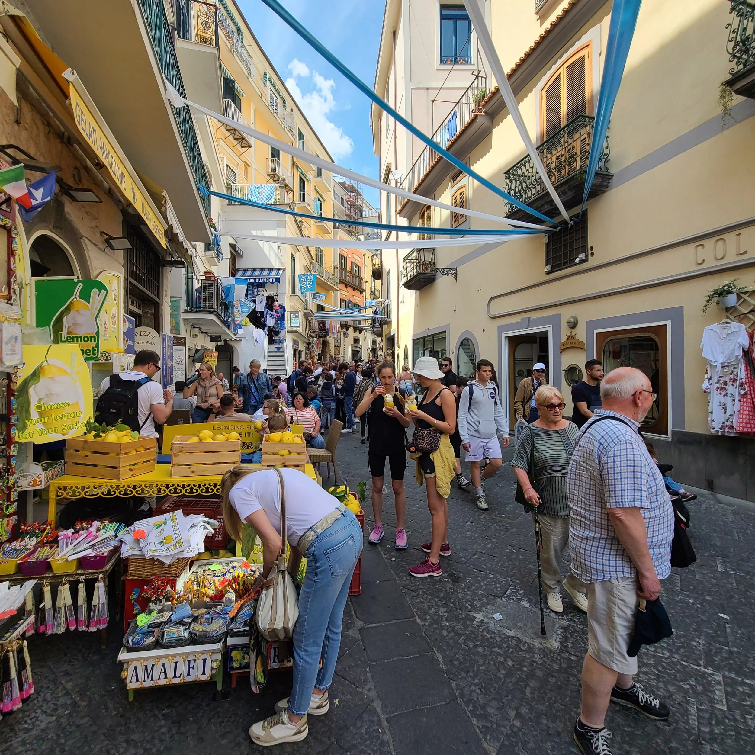 Crowd of people shopping at an outdoor market on a narrow street lined with shops, with colorful signs, clothing, and souvenirs, and buildings with balconies and laundry hanging.