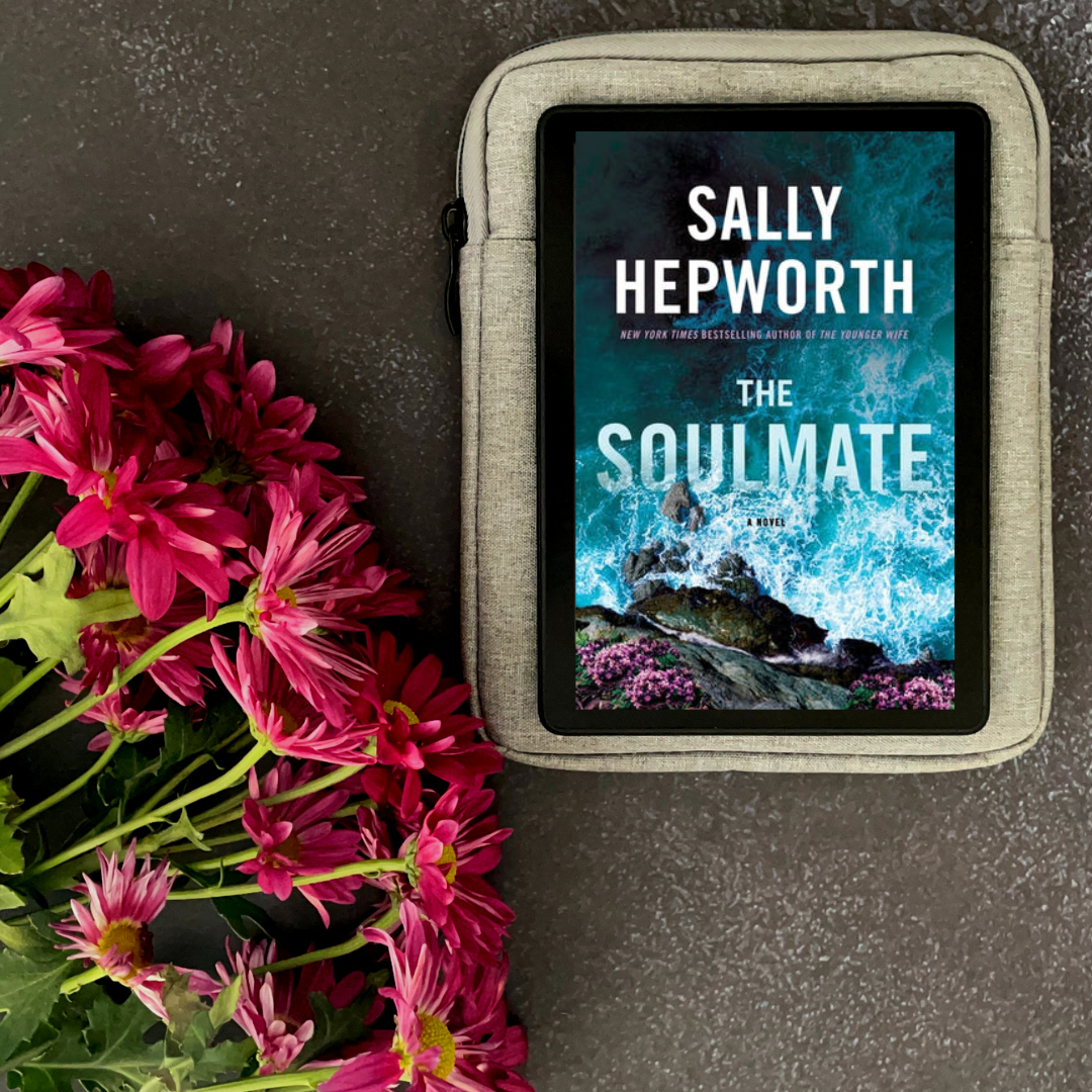 Vibrant pink flowers sit on a dark grey surface next to a kindle displaying the book cover for The Soulmate by Sally Hepworth. The cover shows ocean waves crashing into rocks.