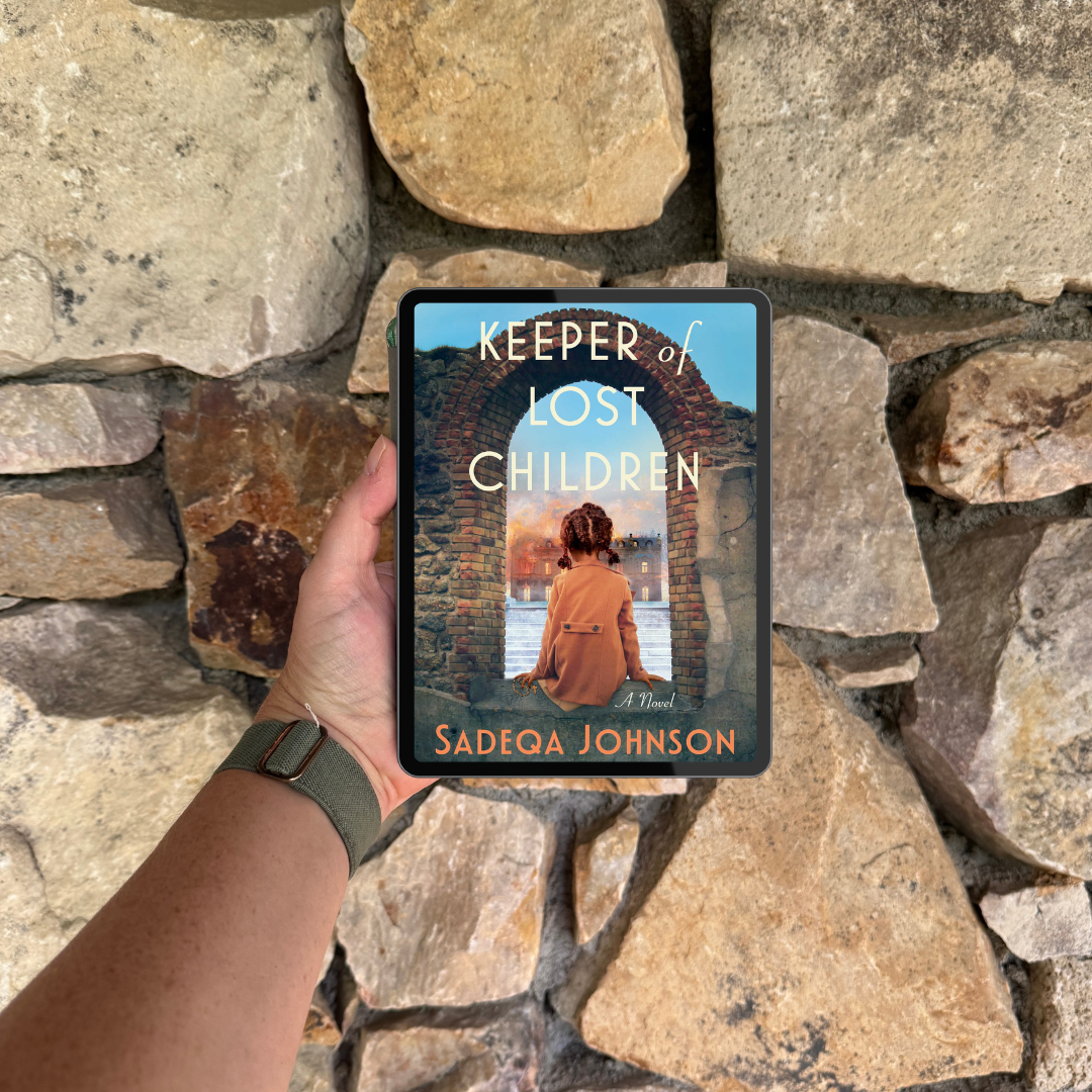 An e-reader displaying the book Keeper of Lost Children by Sadeqa Johnson is held in front of a wall made of large rocks.