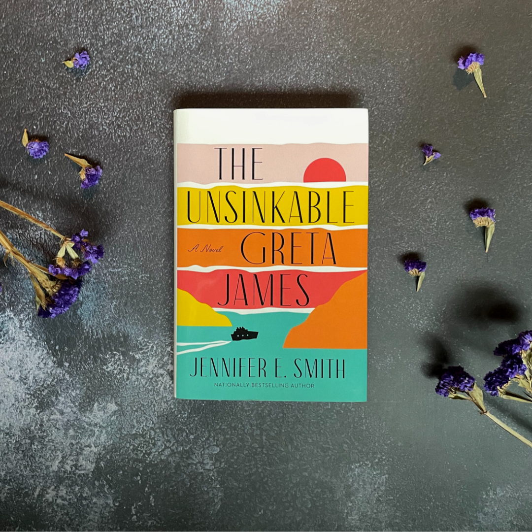 Hardcover book The Unsinkable Greta James by Jennifer E. Smith sits on a dark concrete surface with dried purple flowers scattered.