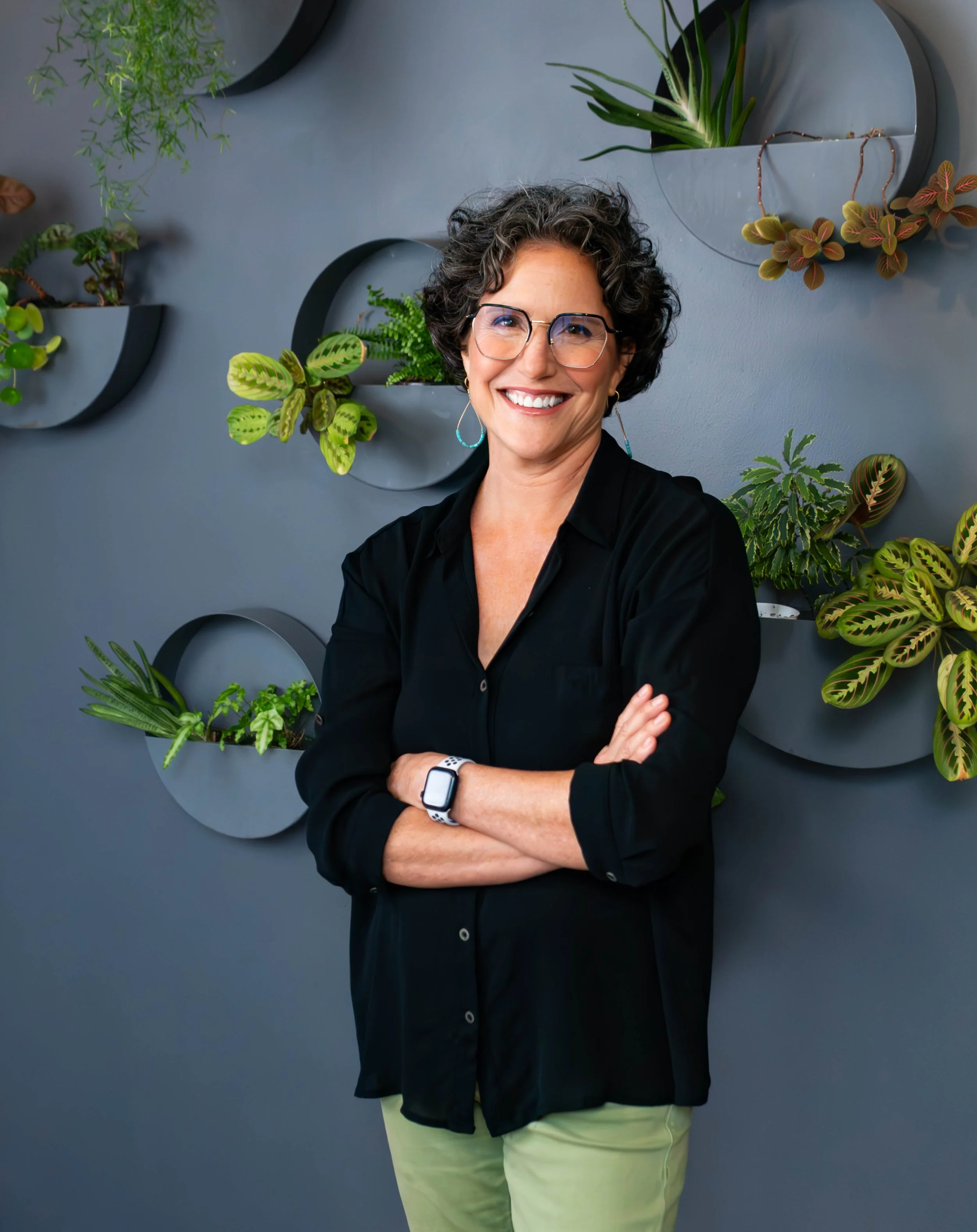 Woman with short curly dark hair wearing glasses, black shirt, and light green pants, smiling with arms crossed, standing in front of a gray wall with mounted decorative planters filled with various green plants.