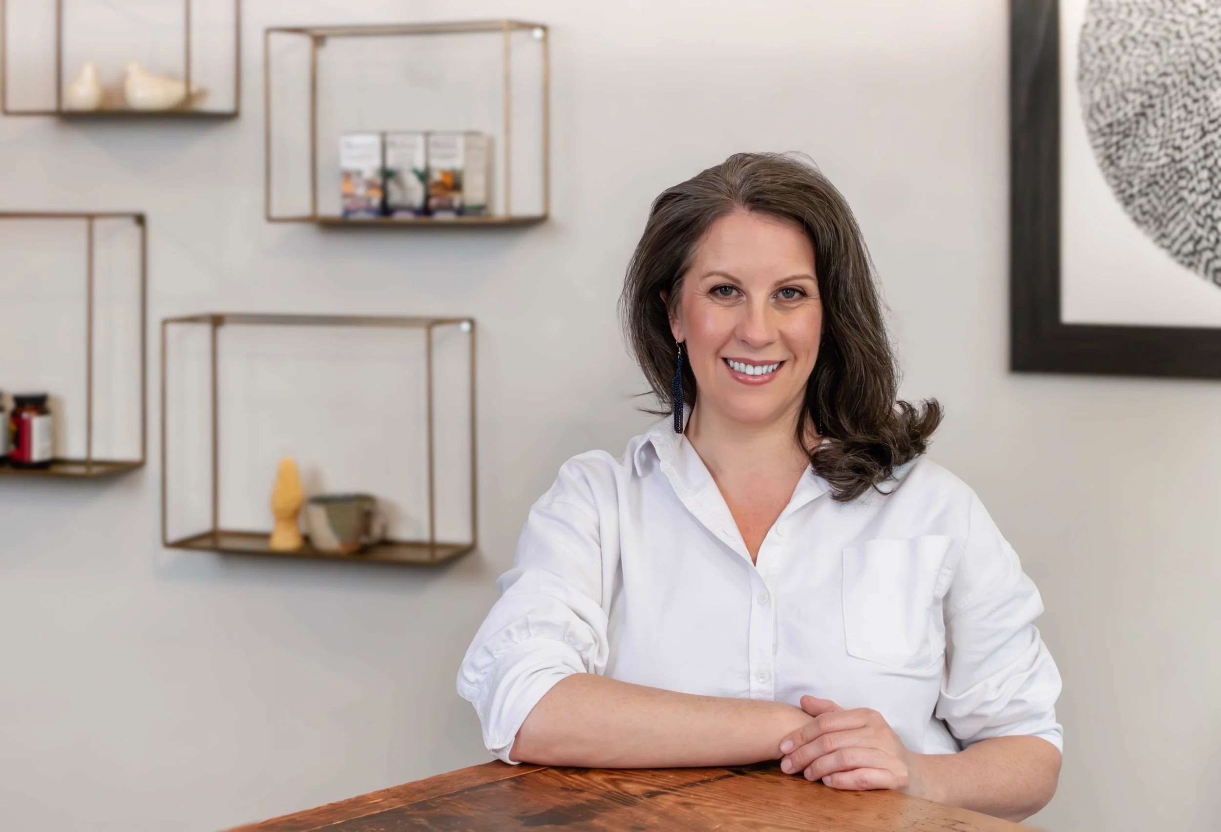 A woman with brown hair, wearing a white shirt, smiling and sitting at a wooden table in a modern, decorated room with empty metal shelves and wall art.
