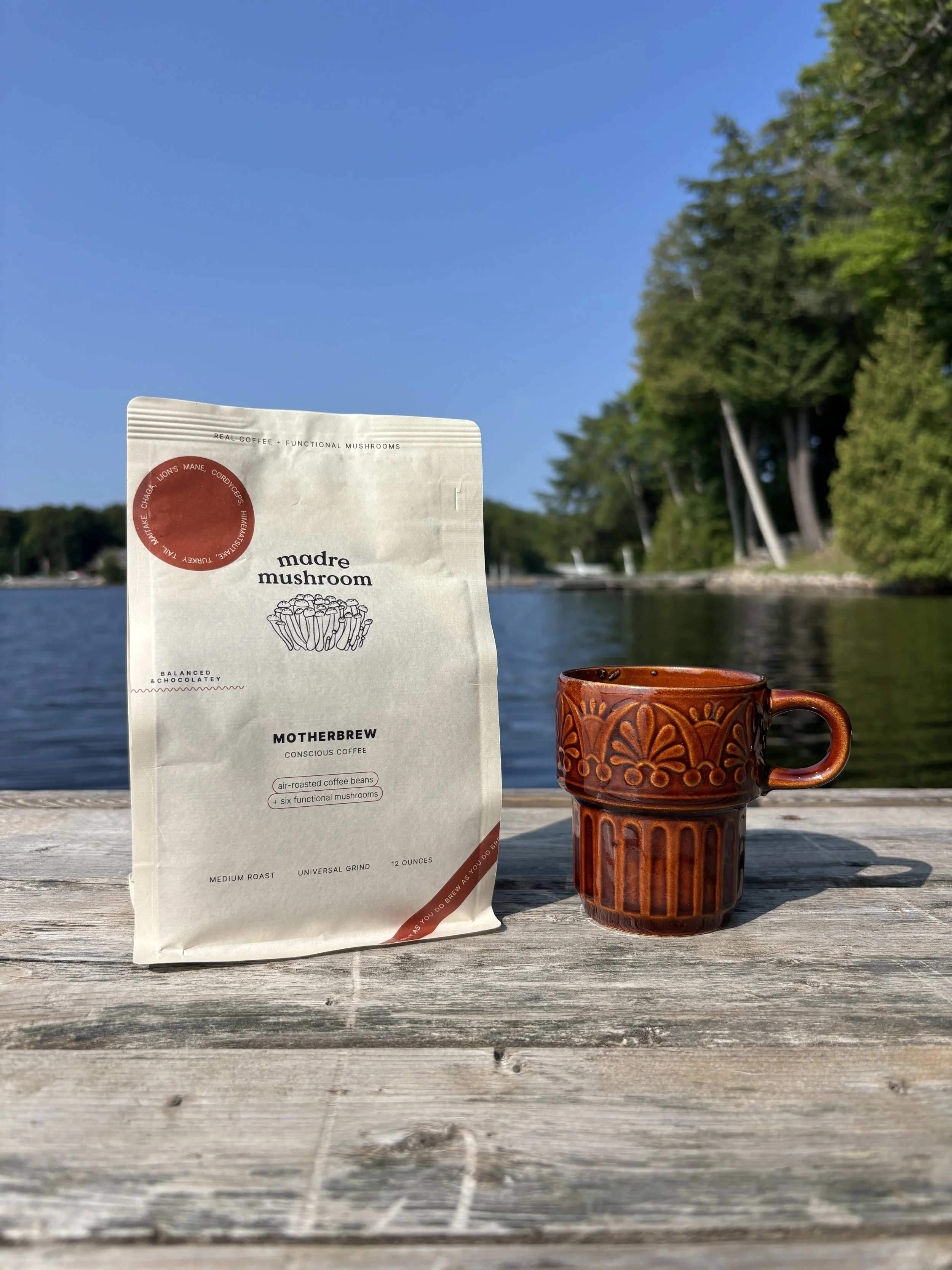 A bag of Madre Mushroom coffee and a decorative ceramic mug on a wooden table outdoors with water, trees, and a blue sky in the background.