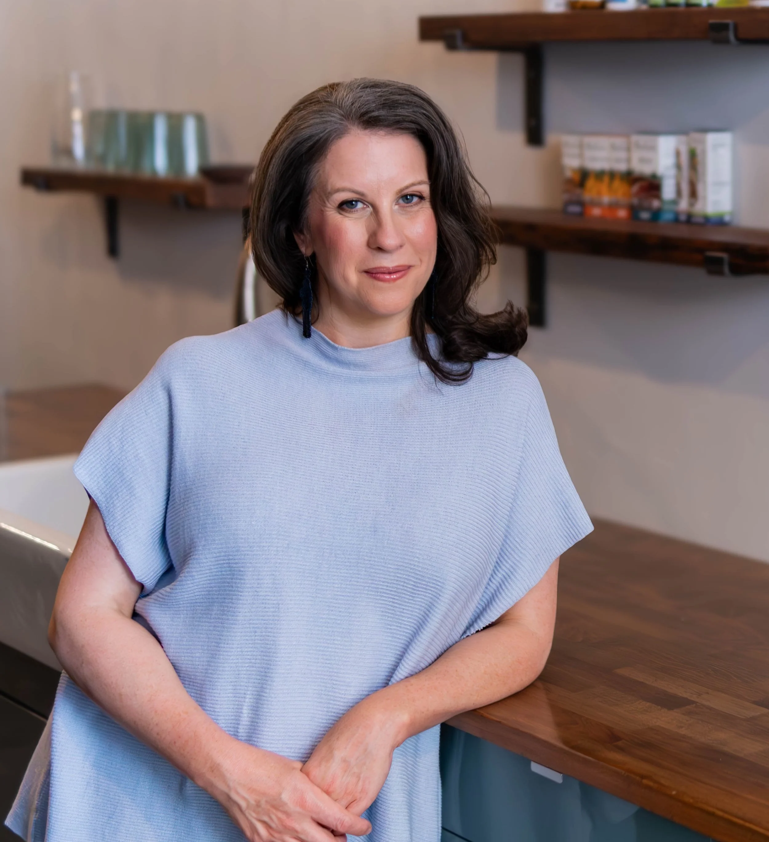 A woman with shoulder-length brown hair and blue eyes wearing a light blue top, standing at a wooden counter in a kitchen or room.