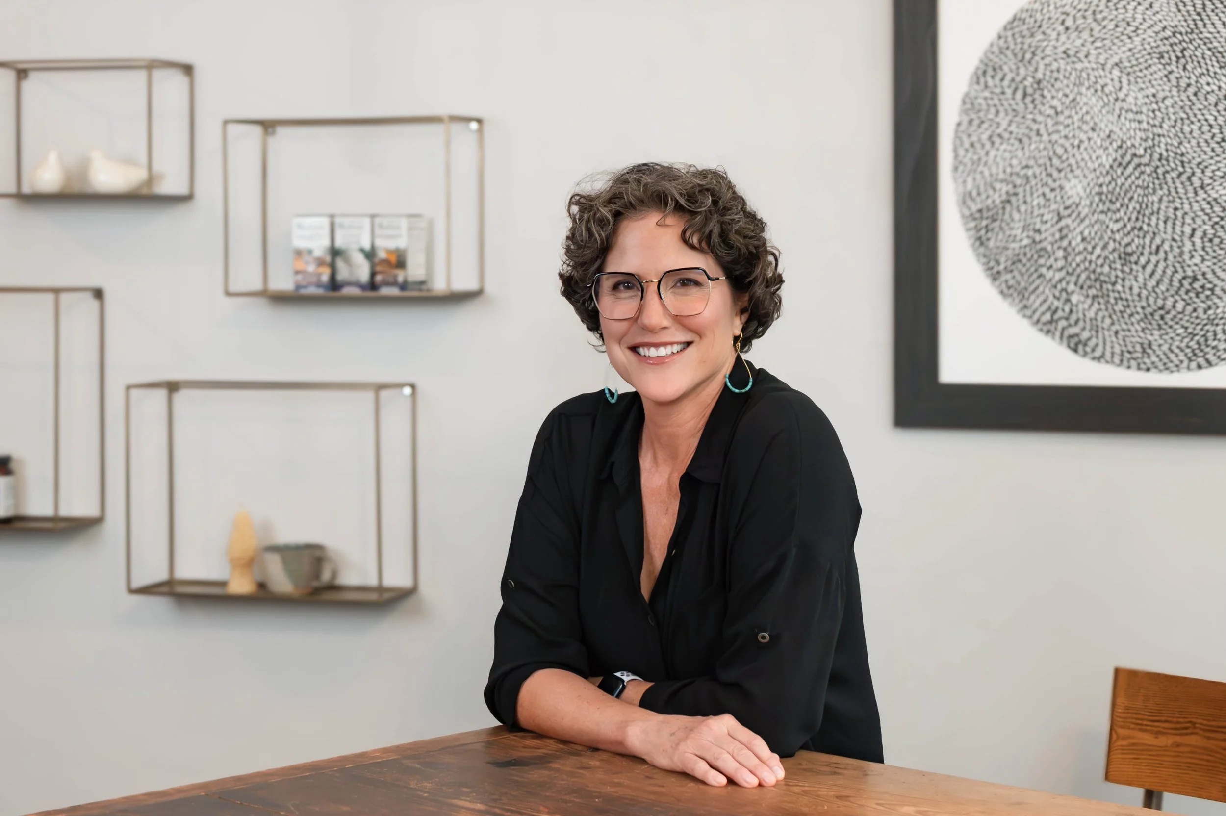 A woman with short curly hair, glasses, and turquoise earrings smiling while sitting at a wooden table in a modern, minimalistic room with wall shelves and abstract art.