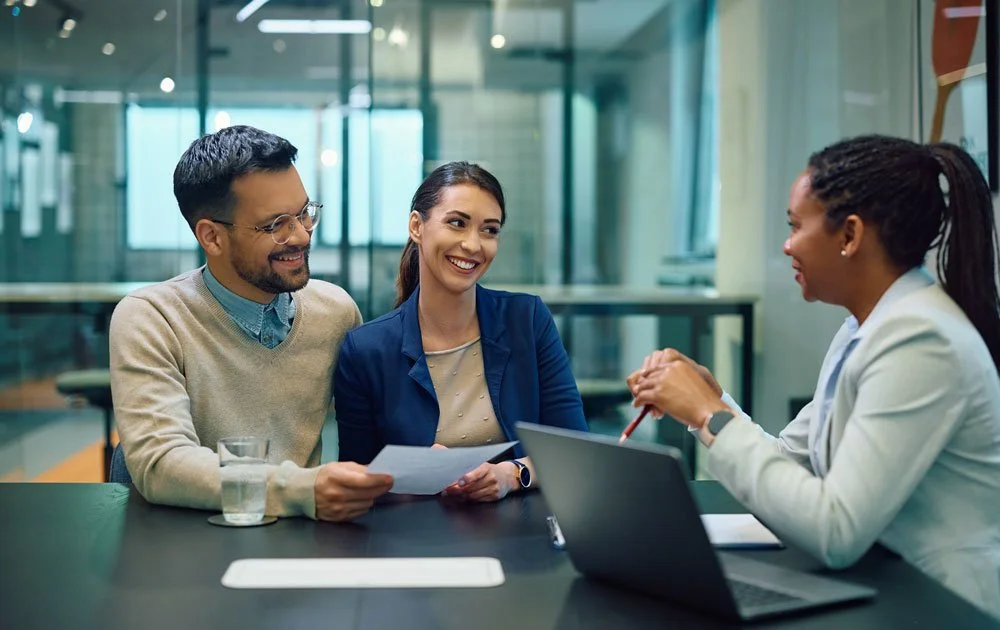 Three people sitting at a table in a modern office, engaged in a friendly discussion. The woman on the right is speaking to a man and a woman on her left, who are listening and smiling. There is a laptop, a glass of water, and papers on the table.