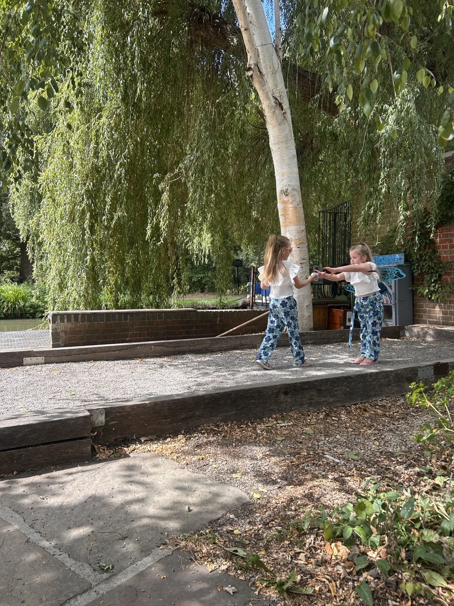 Two young girls with long hair standing on a sidewalk near a tree, exchanging objects outdoors during daytime.