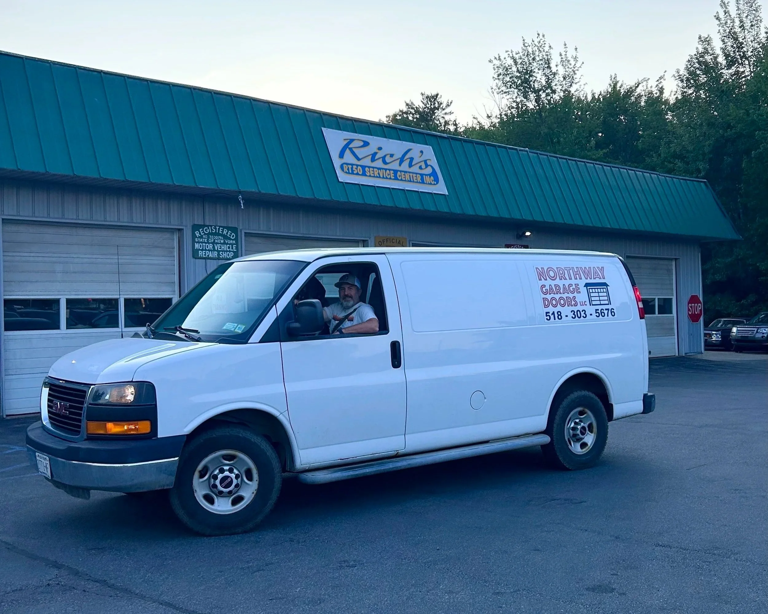 A white GMC cargo van with lettering for Northway Garage Doors parked in front of a garage and auto repair shop called Rich's RTV Service Center Inc. The shop has a blue metal roof and a sign with its name. A man is sitting in the driver's seat.