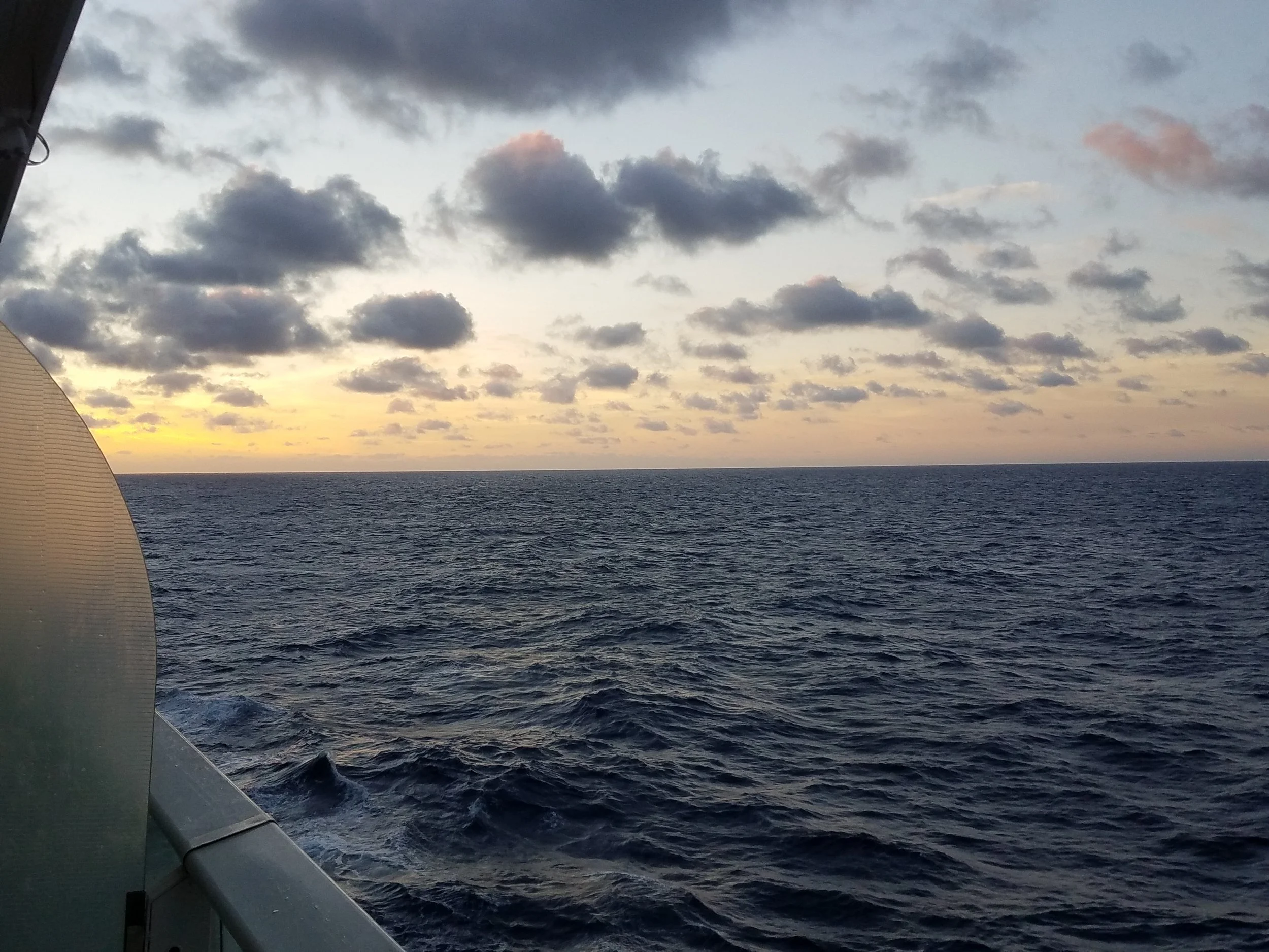 View of the ocean with waves and a partly cloudy sky during sunset or sunrise, seen from a ship or boat.