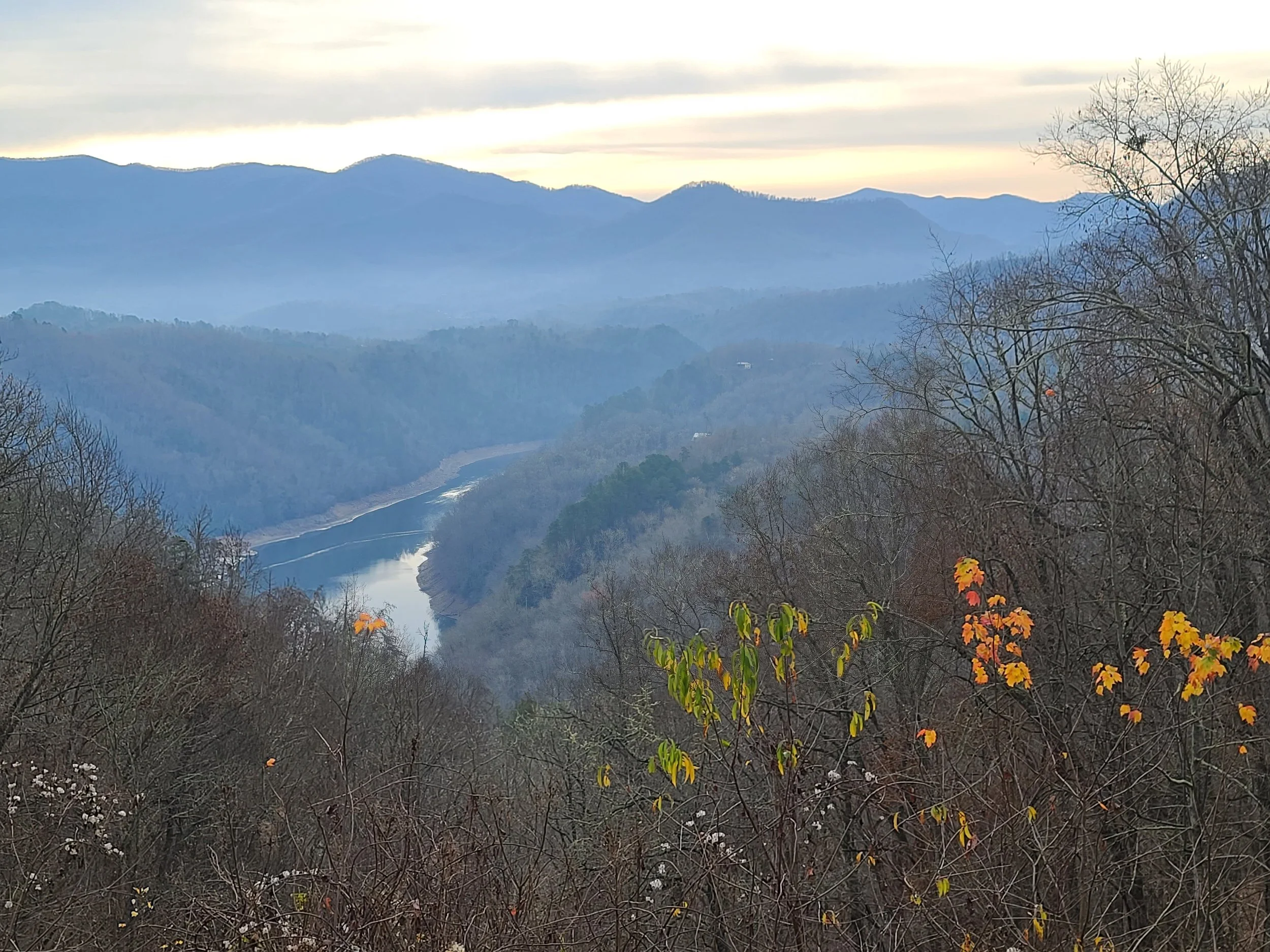 Mountain landscape with layered mountain ranges, a river flowing through the valley, and leafless trees in the foreground, some with orange and yellow leaves.