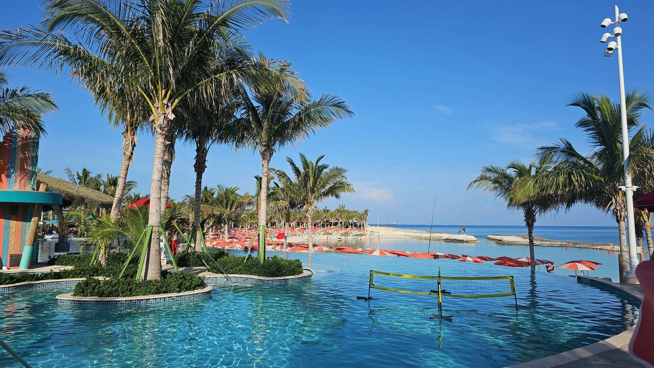 A tropical beach resort with palm trees, a swimming pool, orange umbrellas, and a sandy shoreline with a calm ocean under a blue sky.