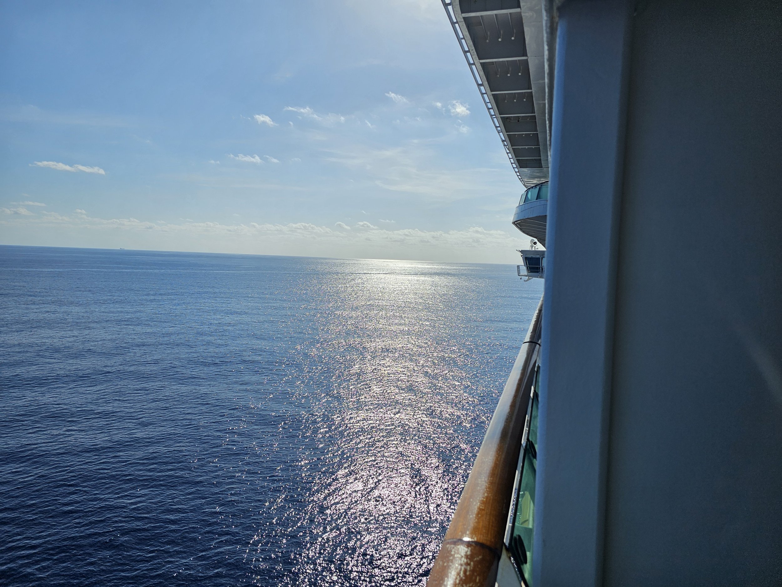 View from a cruise ship balcony showing calm ocean waters with the sun reflecting on the surface, a clear sky with scattered clouds, and part of the ship's railing and structure on the right side.