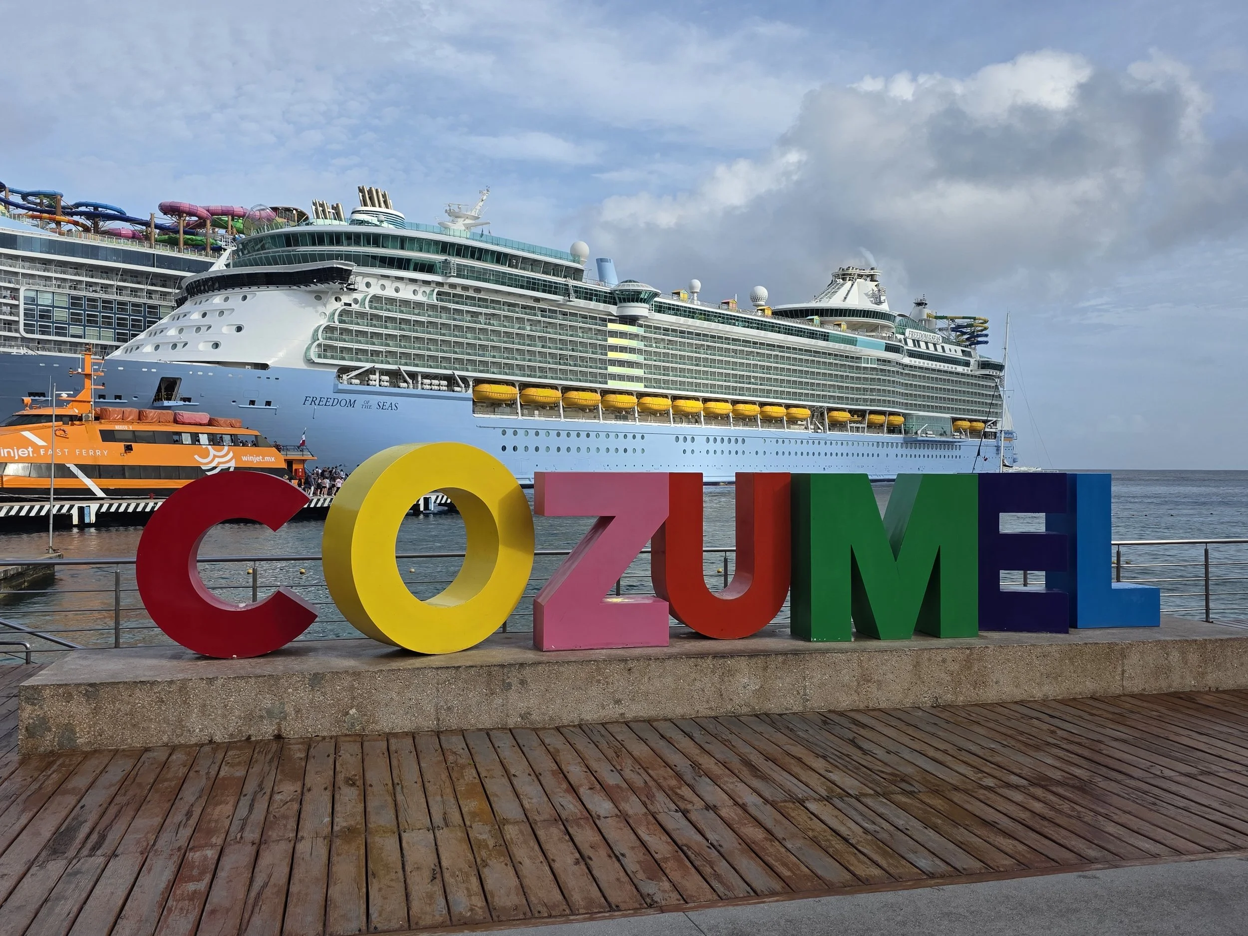 A large cruise ship docked at a port with colorful 'LOVE' letters in the foreground. The cruise ship has multiple decks, water slides on top, and the words 'FREEDOM of the SEAS' on its side. There is also a small orange ferry near the ship.