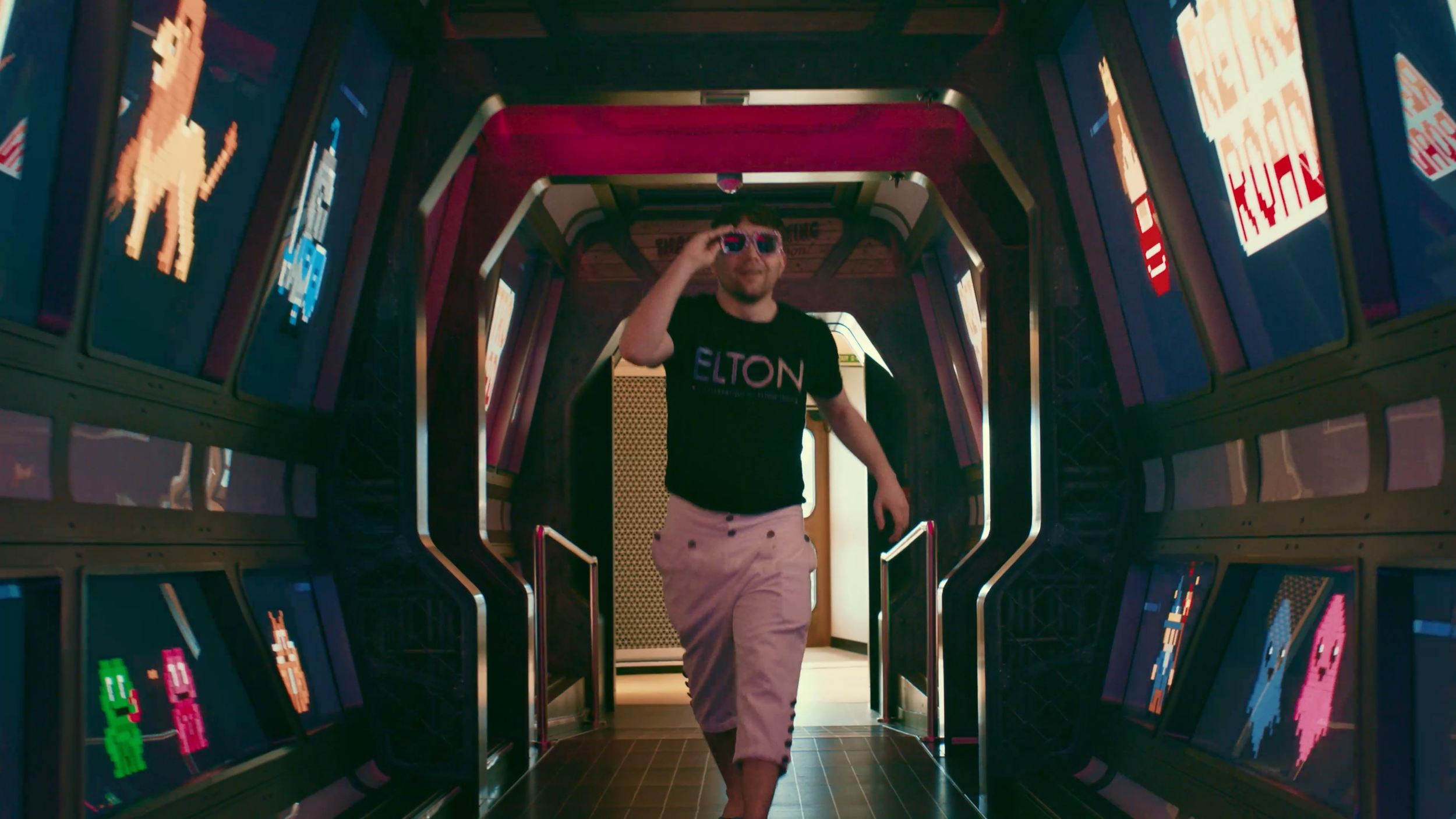 man in black t shirt reading "ELTON" and white pants walking through a hallway full of video screens