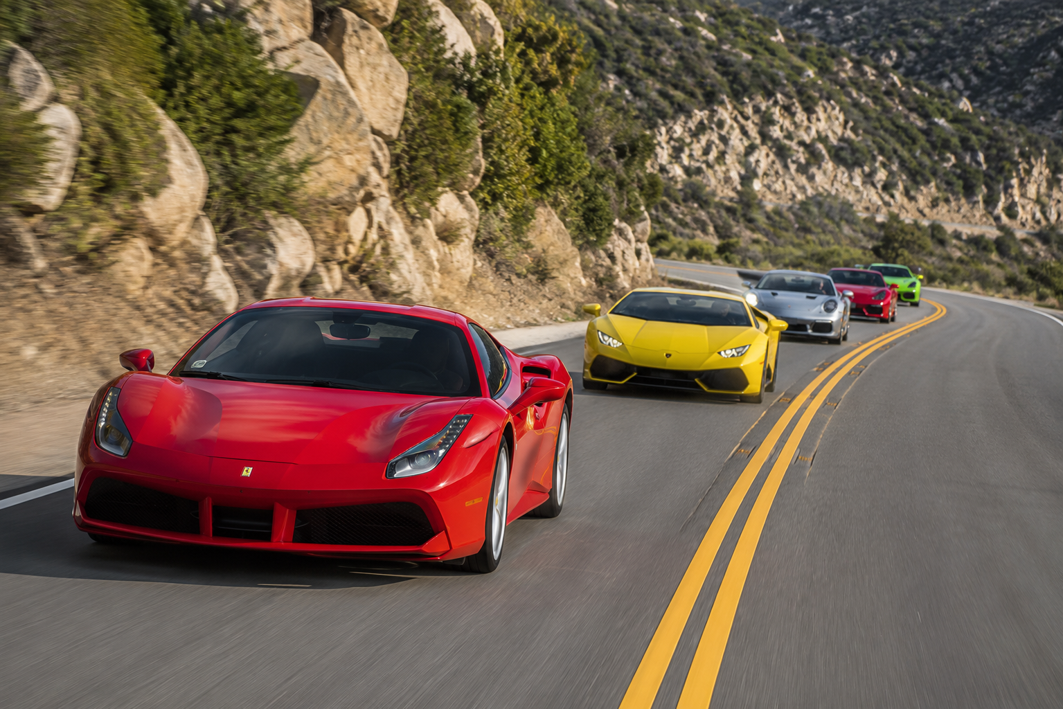 Five luxury sports cars racing on a winding mountain road, including a red Ferrari, a yellow Lamborghini, a silver Porsche, a red McLaren, and a green Lamborghini, with rocky and green hillside surroundings.