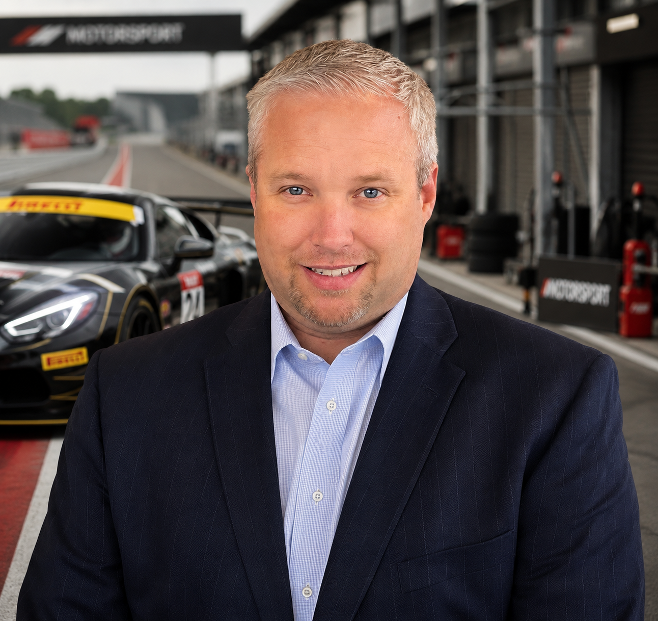 A man in a navy suit and light blue shirt standing in a racing pit lane with a black race car behind him, Ferrari branding visible in the background.