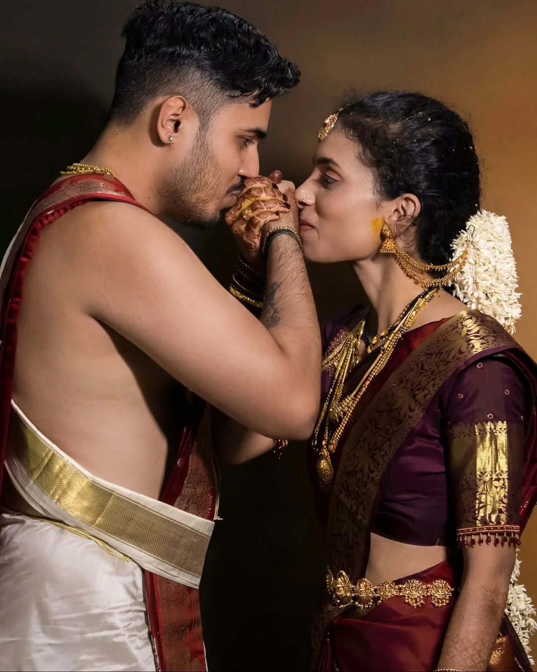 A young man and woman dressed in traditional Indian attire are touching foreheads and holding hands, with the man wearing a cream dhoti and shawl, and the woman wearing a purple sari with gold jewelry and flowers in her hair, during a ceremonial mome