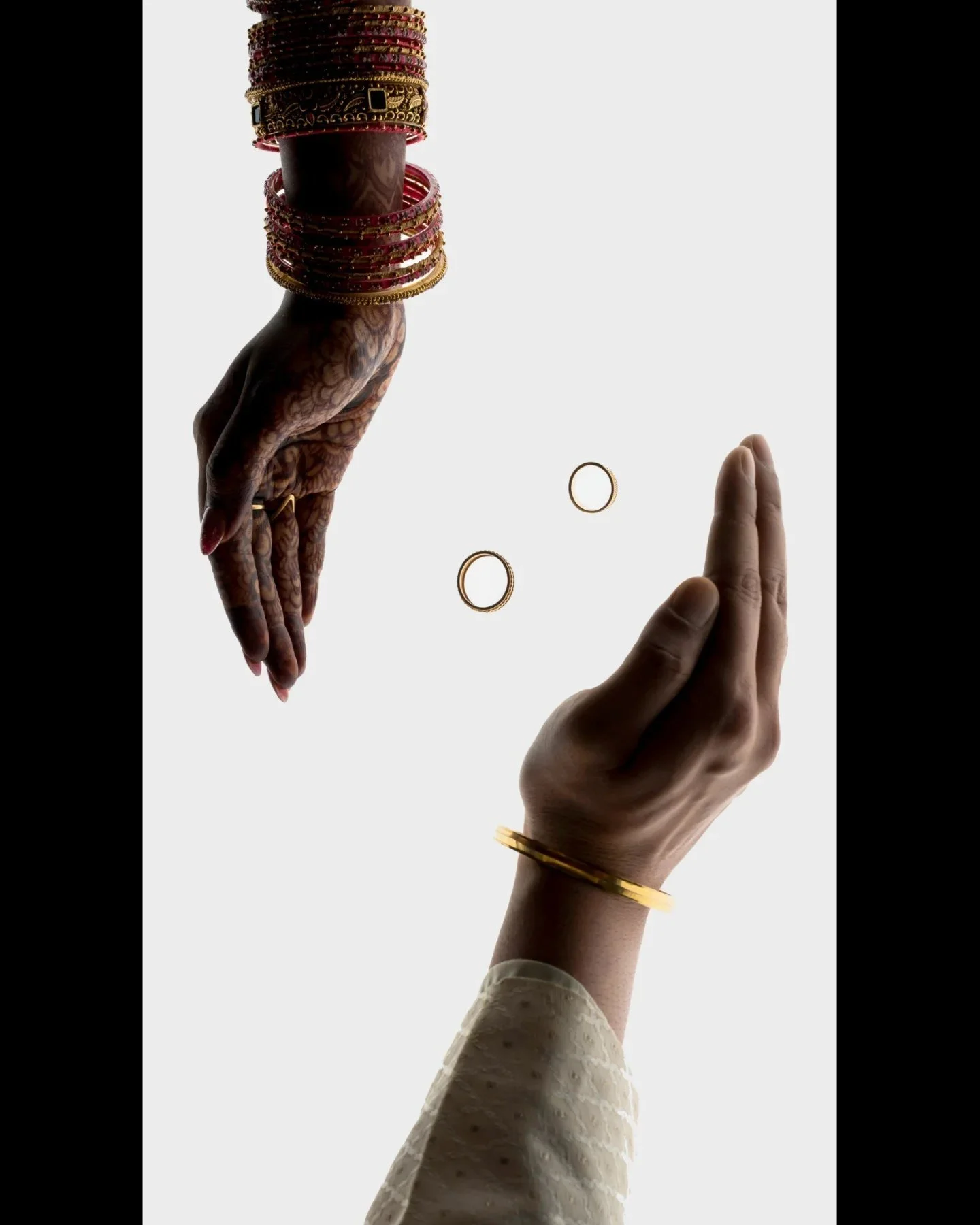 Two hands, one adorned with traditional jewelry and bangles, reaching towards each other with gold rings, against a white background.