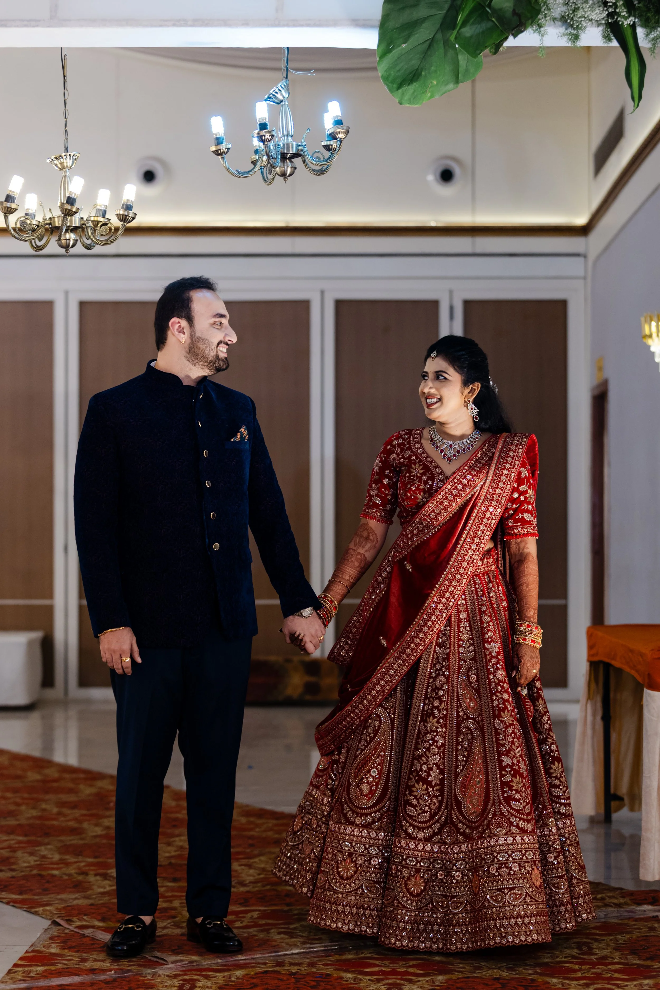 A couple dressed in traditional Indian wedding attire holding hands and smiling at each other in an indoor setting with chandeliers and decorative elements.