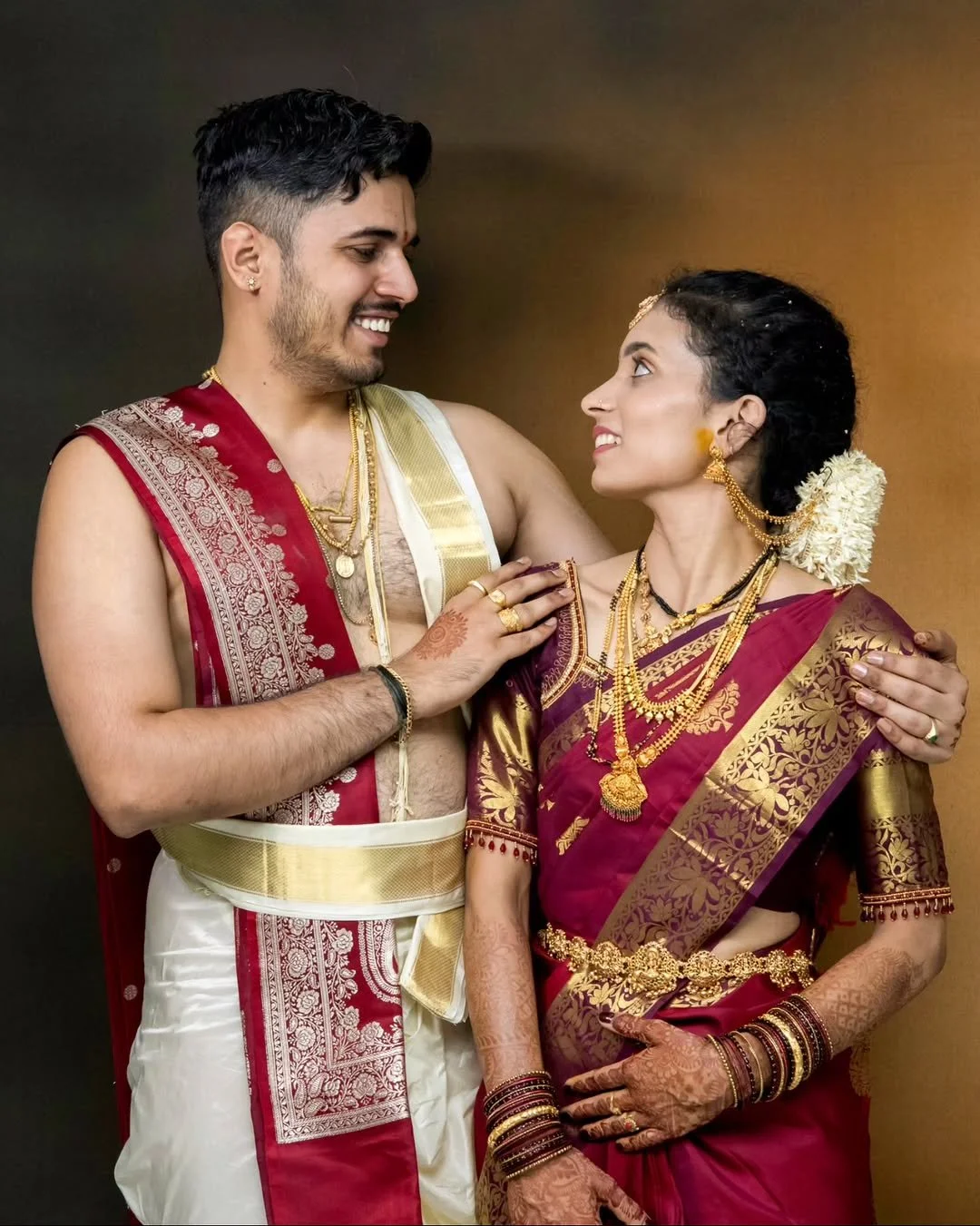 A couple dressed in traditional Indian wedding attire, smiling and looking into each other's eyes, with jewelry, flowers, and henna on their hands, against a warm background.