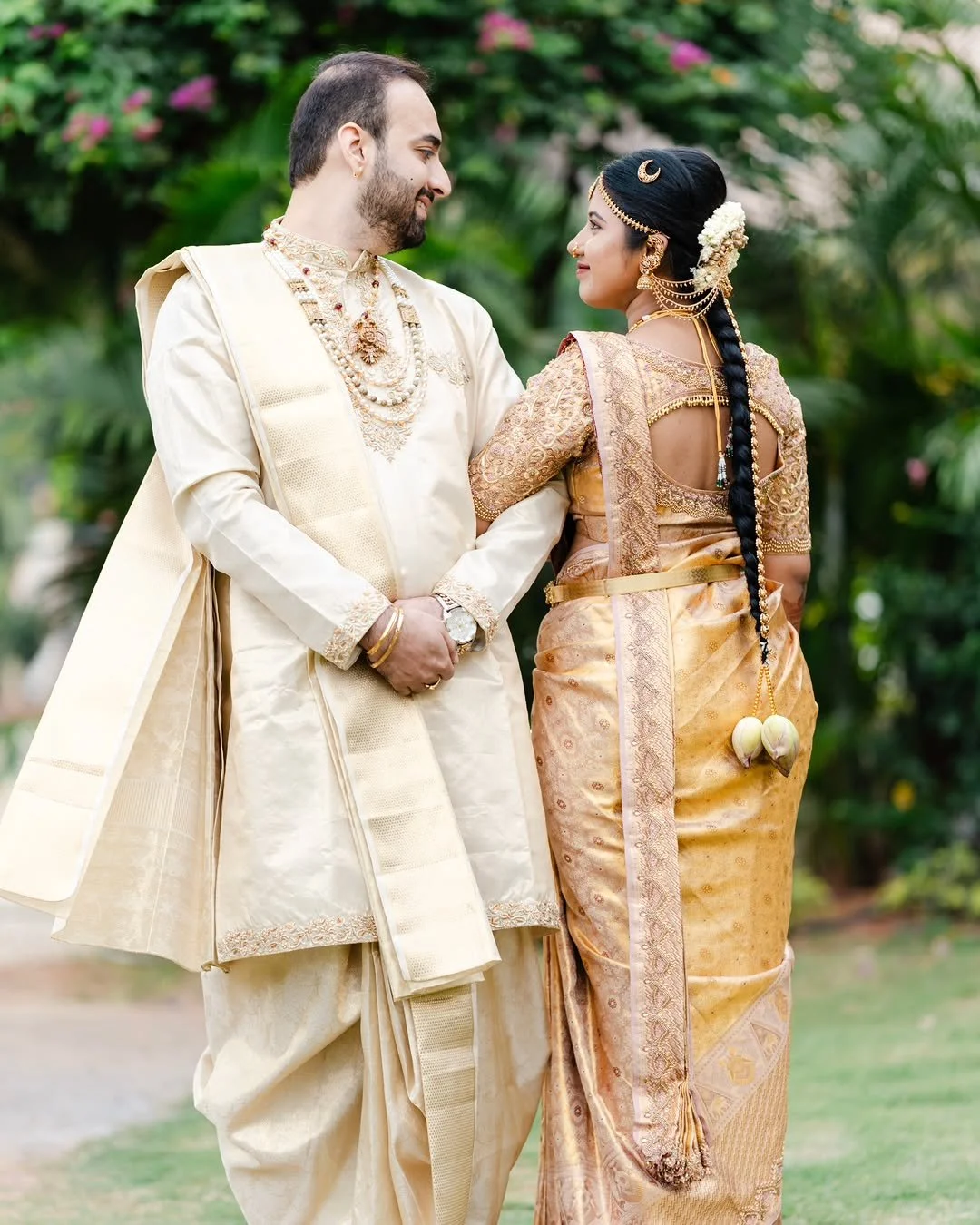 A couple dressed in traditional Indian wedding attire stands outdoors, gazing at each other with greenery and pink flowers in the background.