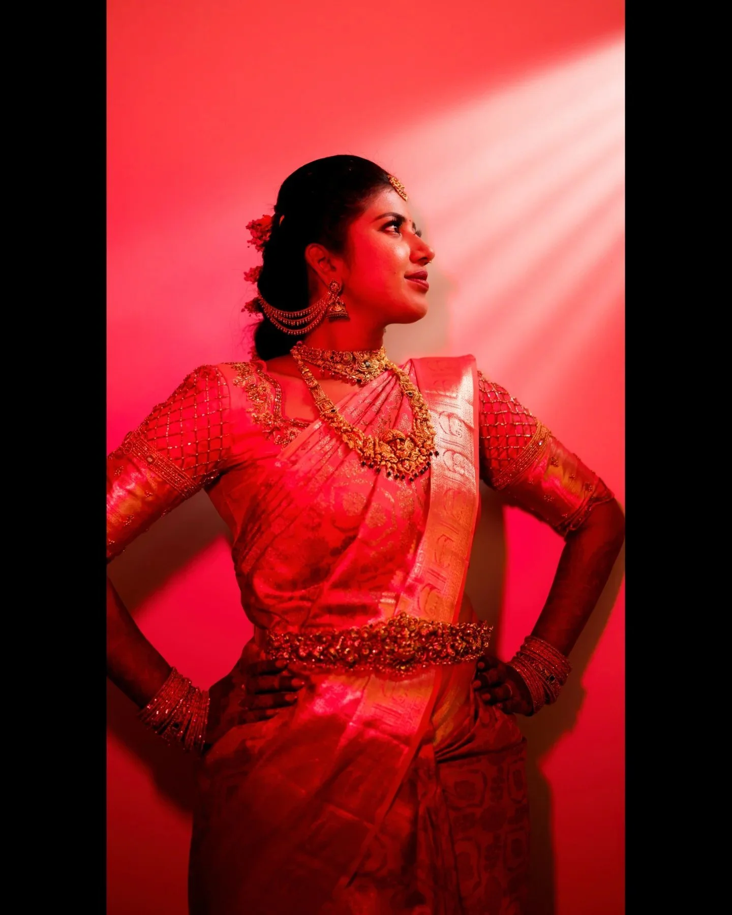A woman in traditional Indian attire, adorned with jewelry, standing in a confident pose with her hands on her hips, illuminated by pinkish-red lighting.